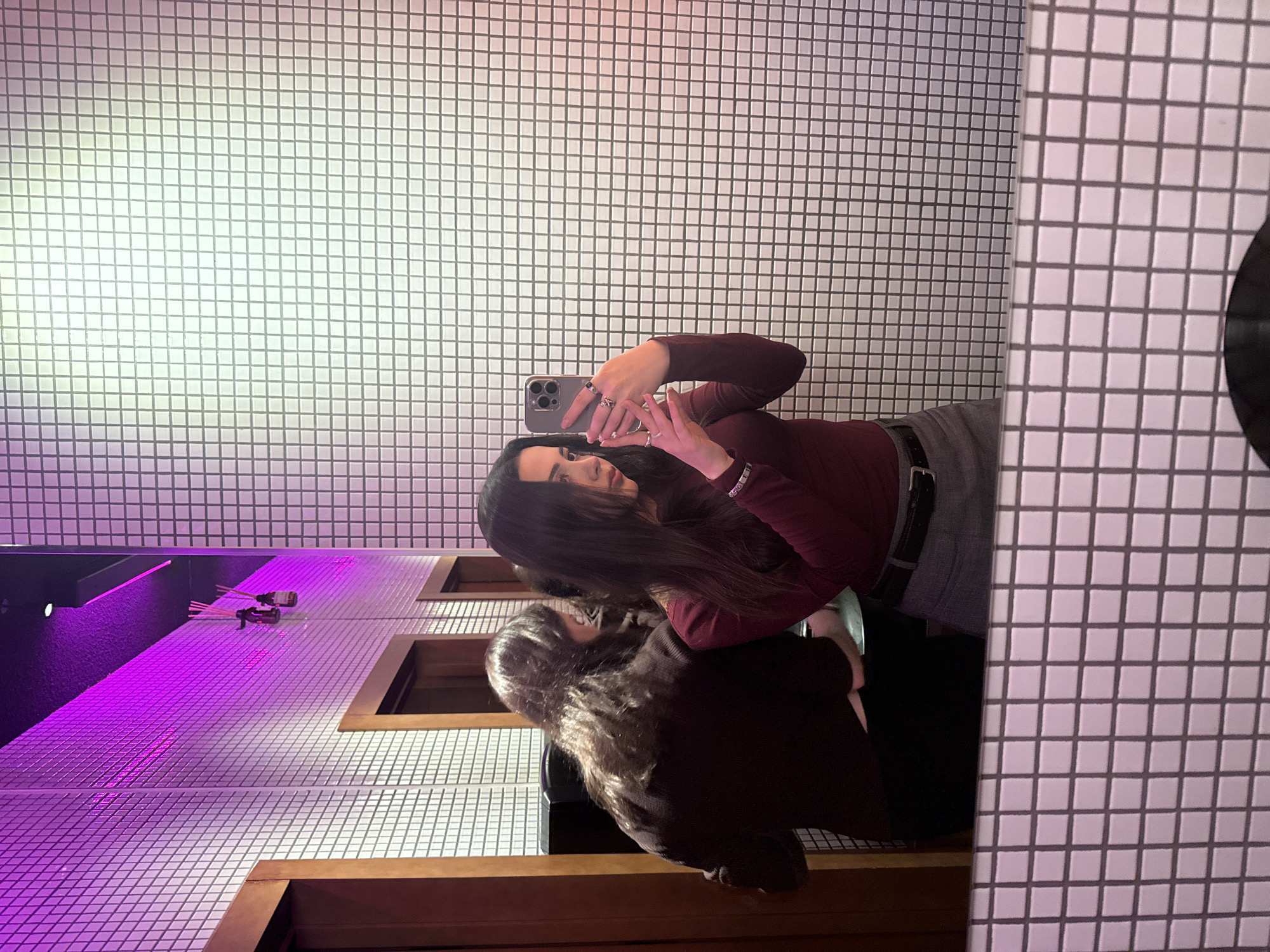 A woman with long dark hair is taking a selfie in a restroom. She is wearing a maroon long-sleeved shirt and a gray skirt with a black belt. She is holding a phone in her hands, focusing on taking the picture. The restroom is decorated with small square white tiles. A portion of a second person, presumably the photographer, is visible in the mirror's reflection. On the left side of the image is a countertop with a basin, illuminated by purple lighting. Two small bottles with sticks in them are on the counter, suggestive of a fragrance diffuser. The restroom appears to be clean and well-lit. There is no information in the image to confirm the specific location in Yerevan, Armenia.