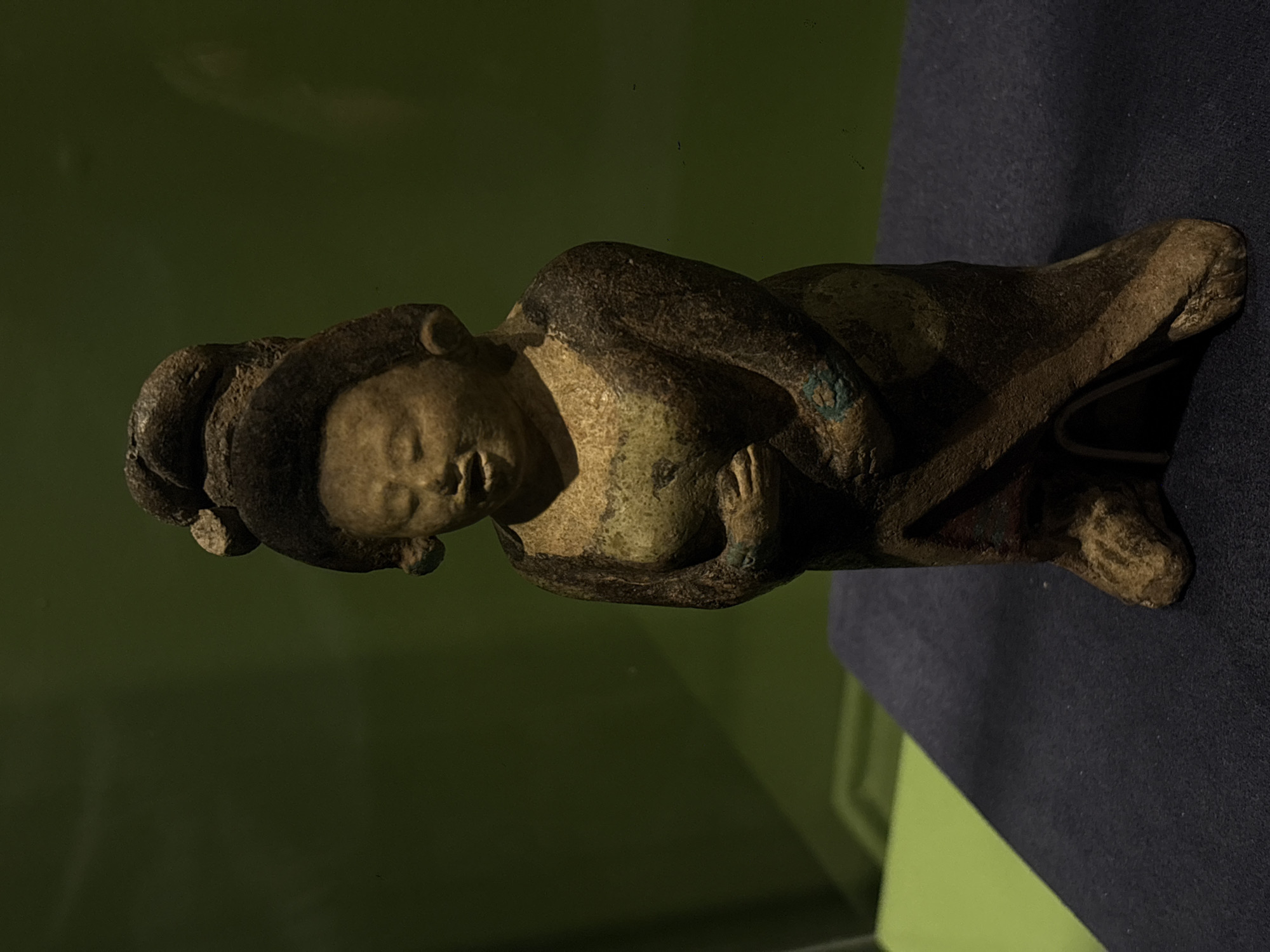 The image depicts a horizontal view of an aged, stone or ceramic sculpture of a reclining female figure. The figure has a ring-like structure encircling the head, possibly a headdress. Her eyes are closed, and her hands are clasped over her chest. The sculpture exhibits discoloration and surface texture variations, suggesting age or exposure to the elements. The figure rests on a dark, possibly velvet surface. The backdrop is a muted green, likely a wall or display case interior. The scene is indoors, presumably within a museum or gallery setting. While the image does not definitively confirm the location as Mexico City, Mexico, the type of artifact suggests a historical or archaeological museum, which could plausibly be located there.