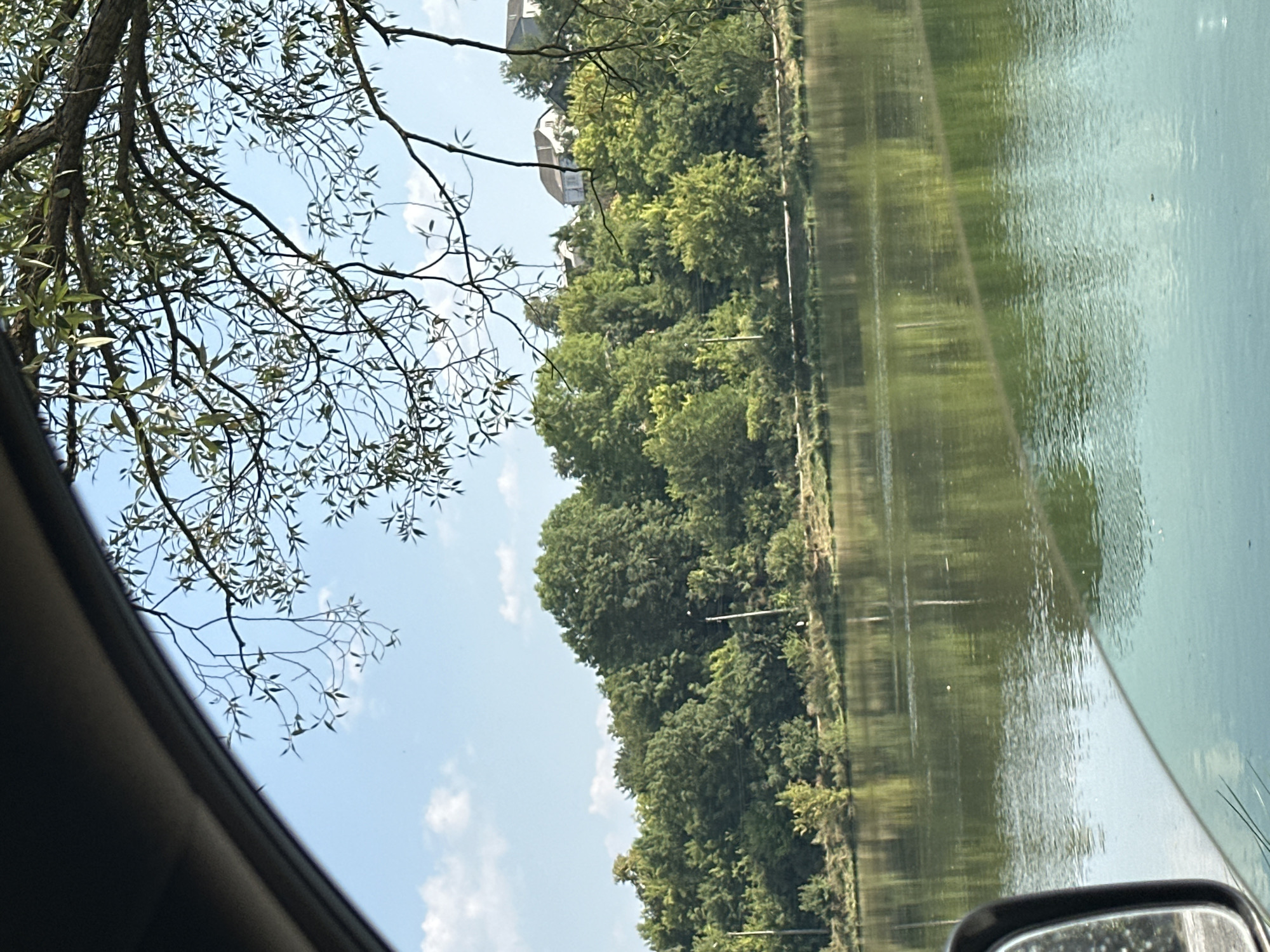 The image shows a view of a body of water, likely a river or lake, from inside a vehicle. The water is calm and reflects the surrounding greenery and sky. On the far bank, a dense line of trees stretches across the horizon, with some houses visible on a slightly elevated area behind the trees. The sky is blue with scattered white clouds. Tree branches, with green leaves, hang into the frame from the top, suggesting the viewpoint is from under a large tree. A car's side mirror and a portion of the car's interior are visible in the lower left corner, framing the scene. The location is identified as Makiv, Ukraine. The scene depicts a natural landscape with man-made structures in the distance, viewed from a personal vehicle.
