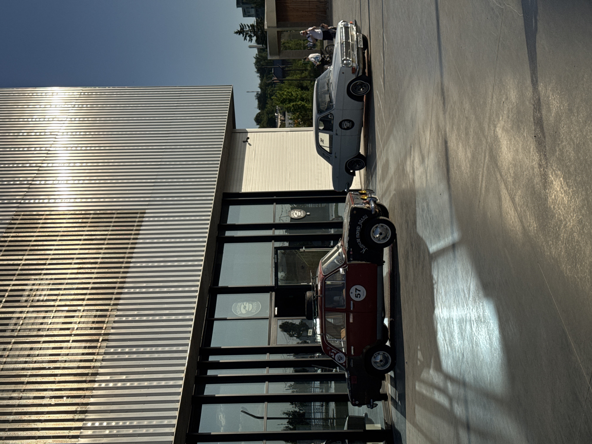 A low-angle, medium shot captures two parked cars against a modern building. The building, featuring horizontal silver stripes, is reflected in the glass-paneled windows. To the right of the red car is a grey car. In the background, some people are visible. The setting appears to be Kazan, Russia.