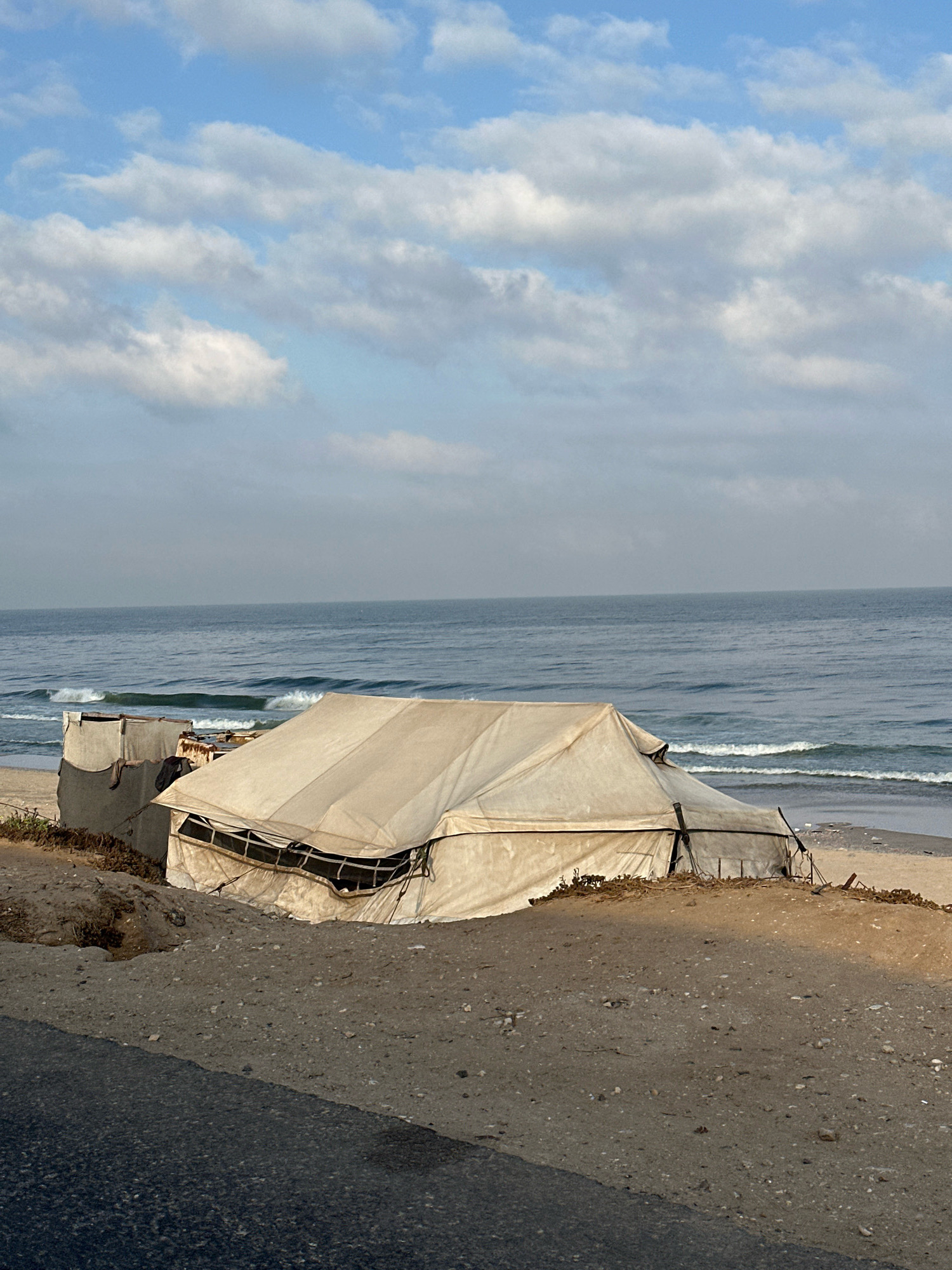 A large, beige tent sits on a sandy incline overlooking the ocean. The tent appears weathered and slightly collapsed in the middle, with metal poles supporting its structure. One side of the tent has a section with visible netting or screening. To the left of the tent, there's a smaller, box-like structure covered with a darker material, possibly a tarp. The ground in the foreground is a mix of sand, gravel, and some sparse, dry vegetation. A dark asphalt road runs along the very bottom of the image, diagonally from the lower left corner. The ocean is visible in the background, with gentle waves breaking near the shore. The sky above is blue with scattered white clouds. No people are present in the image.