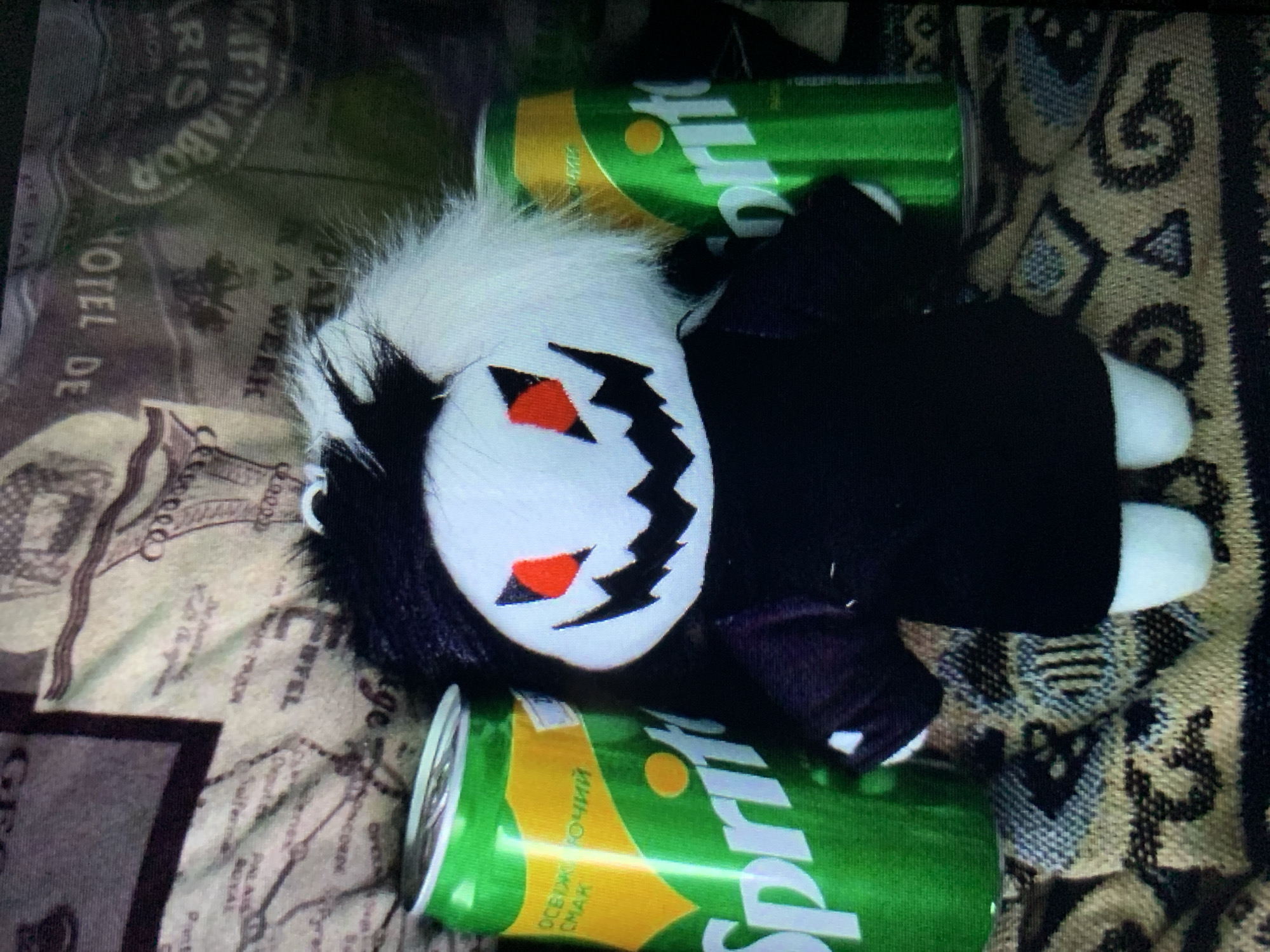 The image depicts a stuffed toy with white and black hair, red eyes, and a black mouth with sharp teeth, dressed in a black outfit. It is positioned between two green Sprite soda cans. The stuffed toy and cans are placed on a patterned fabric surface, which appears to be a blanket or upholstery. The background shows a blurred image of a fabric with text, possibly indicating a map or vintage advertisement. The scene does not provide any specific information about the city of Lymanka, Ukraine, or any forensic details that can be extracted about its location there.