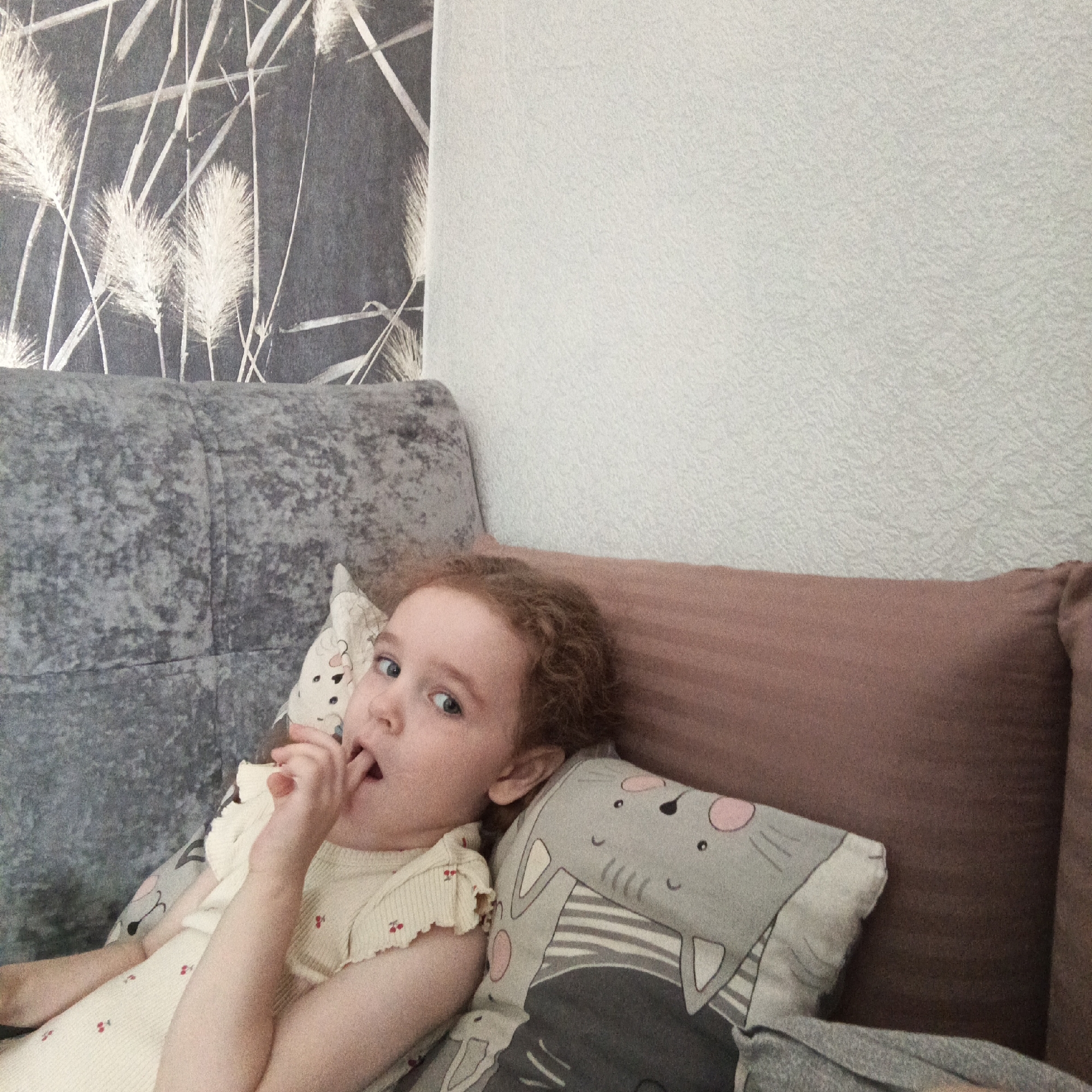 A young girl with curly red hair is lying on a couch, with her index finger in her mouth. She is wearing a light-colored dress with small red cherry patterns. She is surrounded by pillows: one with a cat design and another that is brown. The couch has a gray headrest. The wall behind the couch has a textured pattern on the upper right and a gray wallpaper with white grass-like designs on the upper left. The picture was taken in Марковский, Russia.