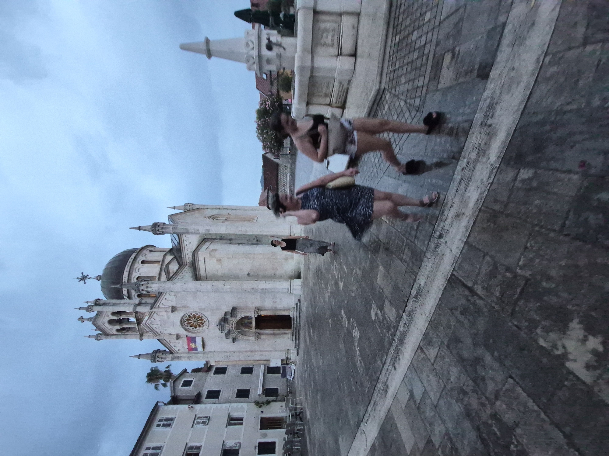 The image depicts an outdoor scene in what appears to be a European town, specifically Herceg Novi, Montenegro. The photograph is oriented vertically, with the sky occupying the upper left portion and a cobblestone plaza dominating the foreground and right side. A large, ornate white church with a dome and intricate stonework stands prominently on the left, dwarfing the surrounding buildings.
In the plaza, three individuals are visible, appearing to be in motion. Two women are walking or posing, one in a dark patterned dress and the other in a light-colored top and patterned skirt. A third person, partially obscured, is also in the vicinity. The overall atmosphere suggests a leisurely stroll or a moment captured candidly.
The time of day appears to be late afternoon or early evening, given the soft, diffused light and the overcast sky. The weather seems mild, as the individuals are dressed in light clothing. The architectural style of the church and the surrounding buildings, along with the cobblestone paving, are strong indicators of a historical European setting.
A notable detail is the flag displayed on the church, which appears to be a triband with a coat of arms. The image is slightly blurry, possibly due to motion or the camera's focus. There is no visible text in the image.