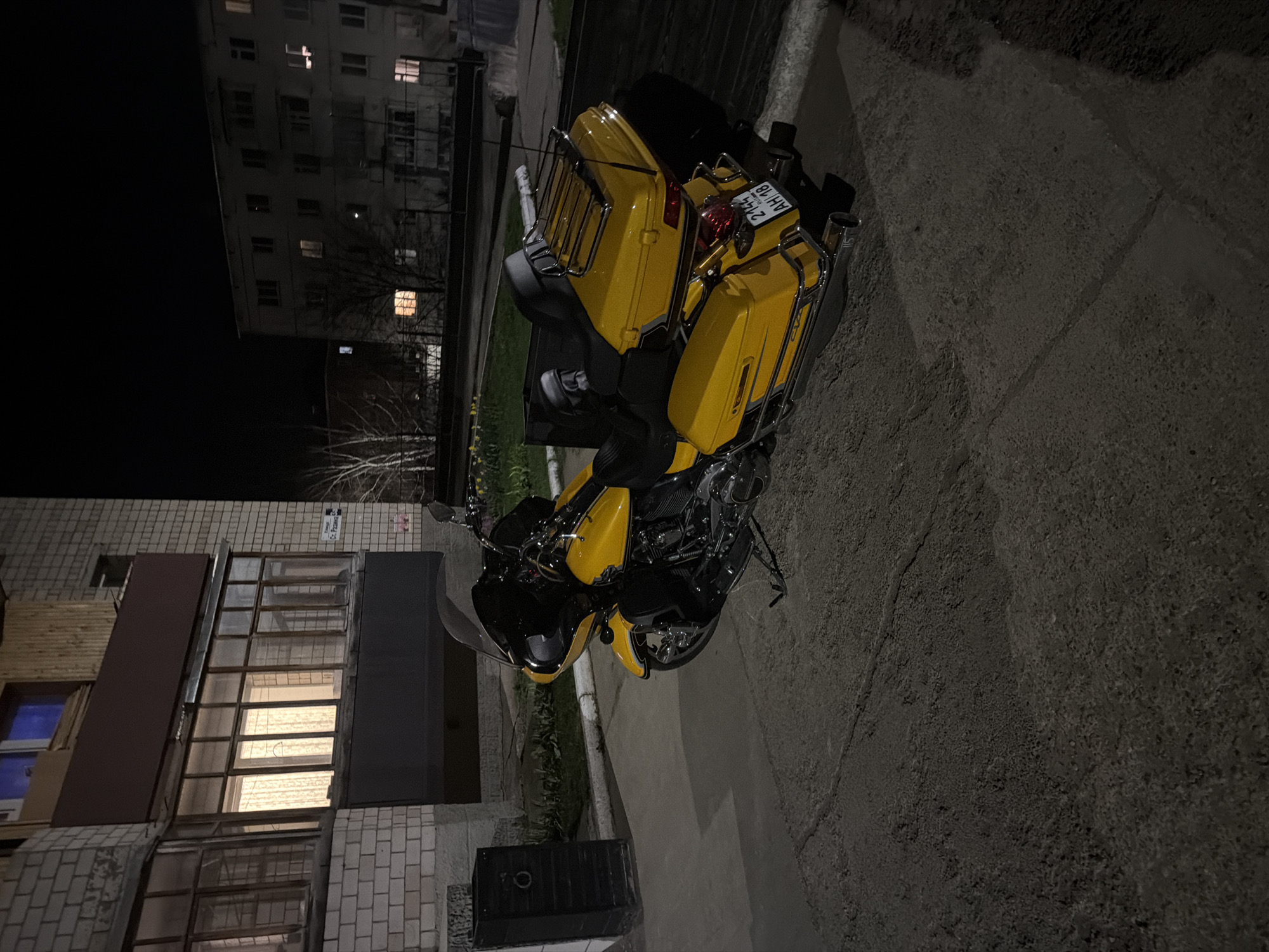 A yellow and black motorcycle is parked on an asphalt surface in Votkinsk, Russia, at night. The motorcycle is a touring model with chrome accents, panniers, and a license plate reading "2144 AH 18". Buildings surround the motorcycle, including a residential building with multiple windows illuminated. The street appears to be deserted, with no people visible in the scene. The lighting is dim, and the overall atmosphere is still.