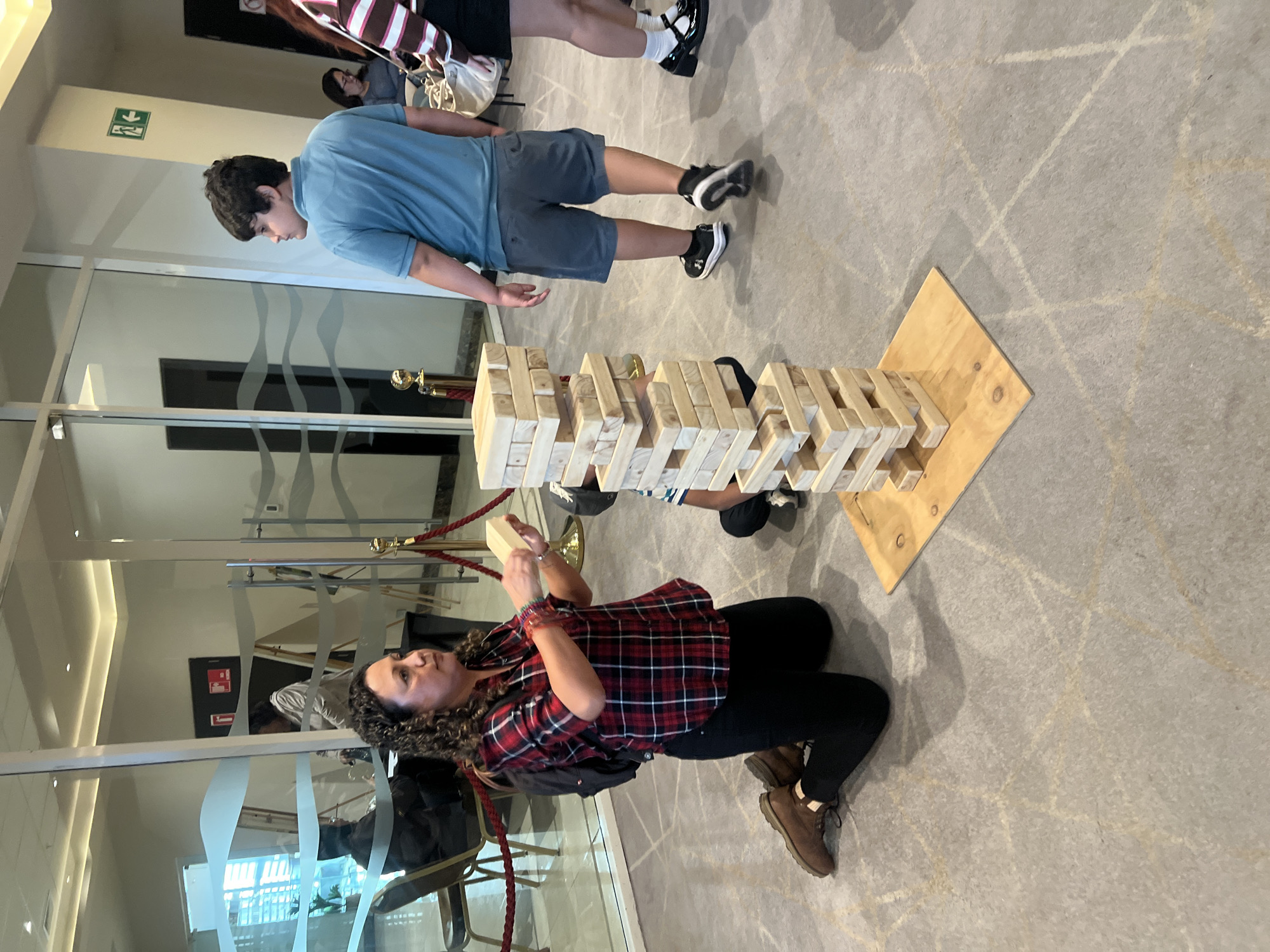 The image shows a group of people playing a game of Jenga indoors. A woman in a red and black plaid shirt and black pants is kneeling on the floor, holding a wooden block from the Jenga tower. A boy in a blue shirt and gray shorts is standing near the tower, observing. Other people are in the background, some are standing and others are walking. The Jenga tower is on a wooden base. The environment is a well-lit indoor space with large windows and a patterned floor. The location of the scene cannot be determined without additional information about the image.