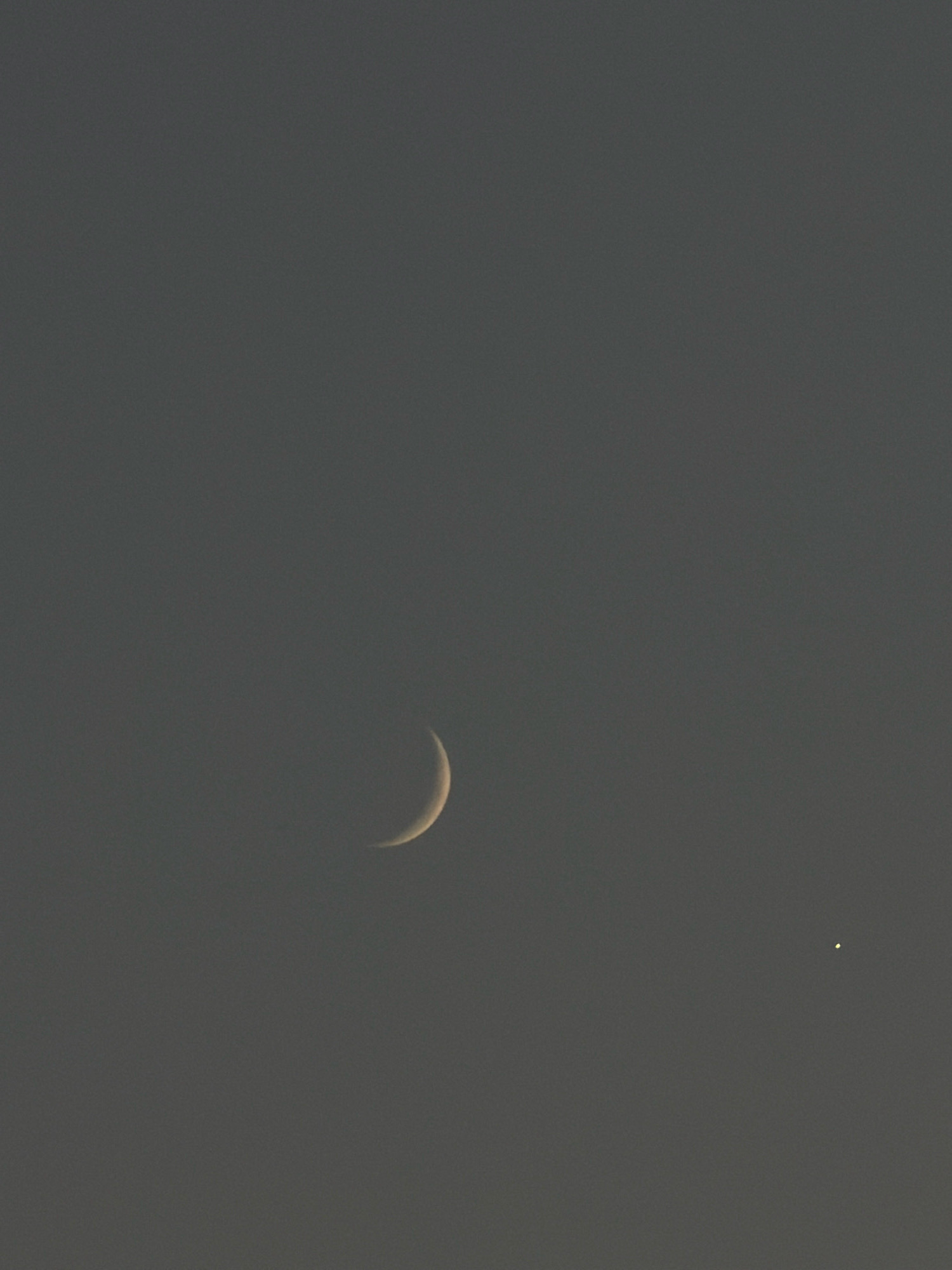 A photograph taken from Sharm El Sheikh, Egypt, shows a crescent moon in a dusky sky, accompanied by a bright star or planet to its right. The moon is positioned in the lower left quadrant of the frame, a thin sliver of light against the muted grey of the atmosphere. The celestial body on the right is a small, bright point, appearing to the right and slightly below the moon. The background is uniformly grey, suggesting either twilight or overcast conditions. There are no visible people, structures, or other objects in the frame.
