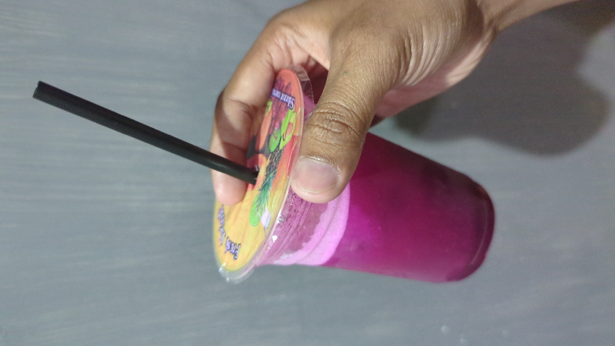 A person's hand is holding a clear plastic cup filled with a vibrant pink beverage. The cup has a clear plastic lid with a colorful graphic design featuring various fruits. A black plastic straw is inserted through the lid, extending upwards. The background is a plain, light gray wall. The image appears to be taken indoors, possibly in a residential or commercial setting within Penjor, Indonesia, given the context of the request.