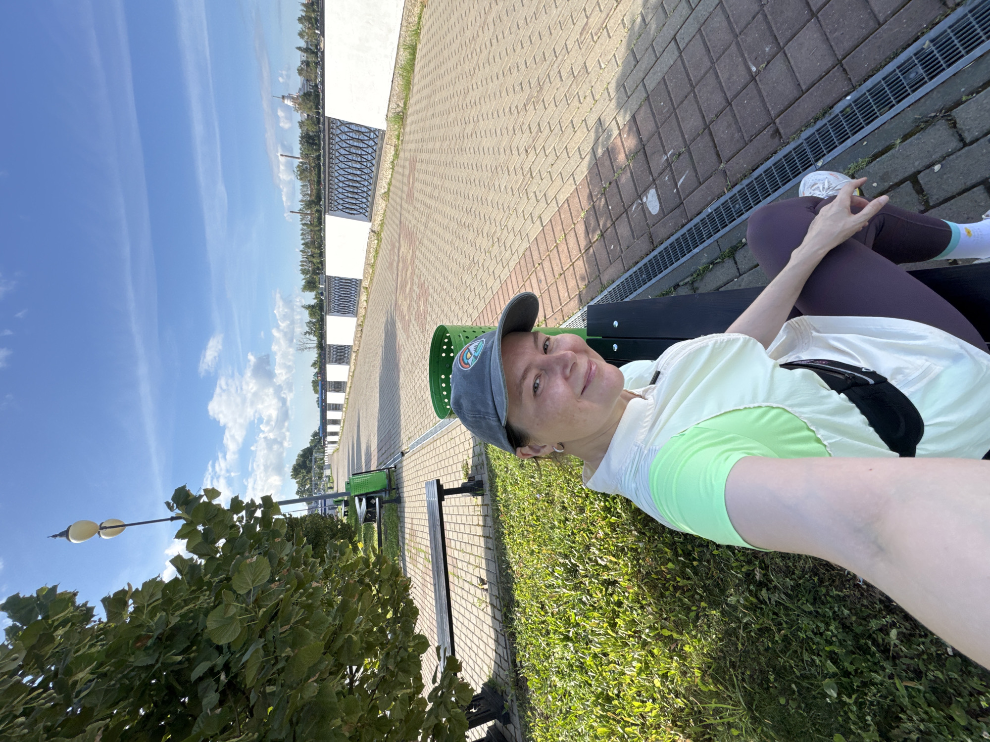 In Izhevsk, Russia, a woman sits on a park bench on a sunny day, with a paved walkway and trees beside her. She is wearing a gray baseball cap and a light-colored shirt with bright green sleeves, as well as dark leggings and white sneakers. A black crossbody bag is visible. The background shows a white railing along a body of water, with buildings and industrial structures in the distance. The sky is blue with scattered clouds.