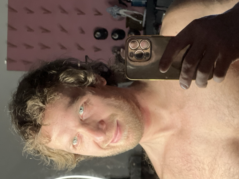 An adult male is depicted in a close-up indoor selfie, captured in what appears to be a mirror reflection. The individual has light skin, light brown curly hair, and a light-colored beard/stubble. His eyes are a striking light blue-green, and he displays a subtle smirk or smile. He appears shirtless, exposing his upper chest and shoulders. A faint reddish mark is visible on the front of his neck. His right hand holds a reflective, possibly gold-colored smartphone, identified by its multiple rear camera lenses, positioned to frame the image.
The background to the left features a visible circular light source, likely a ring light, reflecting brightly. To the right, a wall painted in a pink or mauve hue is visible, adorned with a regular pattern of small, recessed or protruding V-shaped elements casting shadows. Below this pattern, on a surface, several objects are discernible: two dark, spherical items; a light-colored humanoid figurine in a dynamic pose; a dark potted plant; and a blue spray bottle. The setting is an interior room, likely a personal living space. The specific location within Saint Petersburg, Russia, is not discernible from the visual evidence in the image.