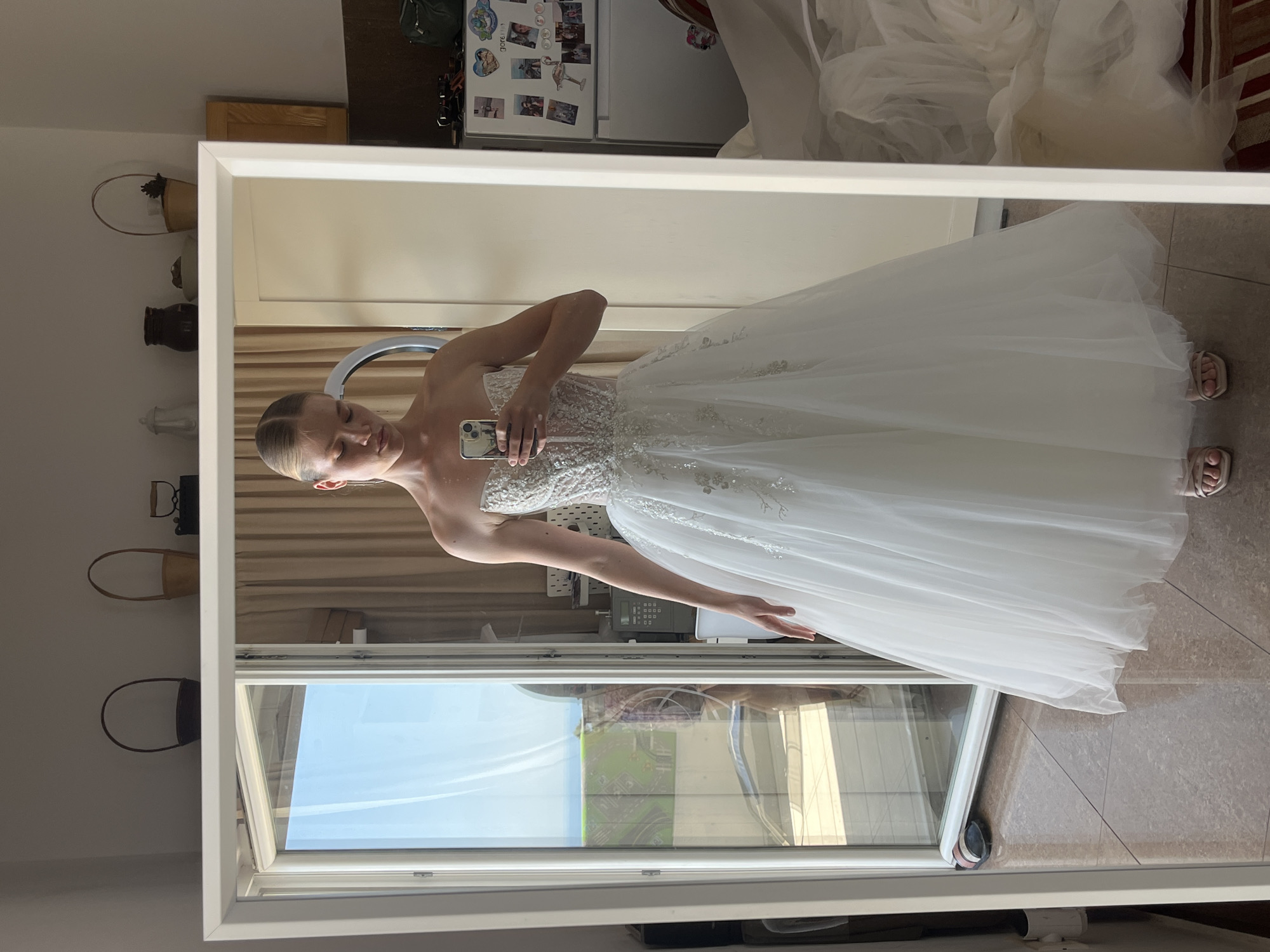 The image shows a young woman standing in front of a full-length mirror, wearing a white strapless wedding dress with a beaded bodice and a full tulle skirt. She is holding a smartphone in her right hand, taking a selfie. The mirror reflects the room, which appears to be a bedroom or dressing area. In the background, there is a large window with a view of the sea and some greenery, suggesting a coastal location. The room contains a refrigerator covered in magnets and a sewing machine. The flooring is tiled. The overall scene conveys a sense of personal preparation for an event, possibly a wedding. Without additional information, the specific location within Budva, Montenegro, cannot be determined from the visual content alone.