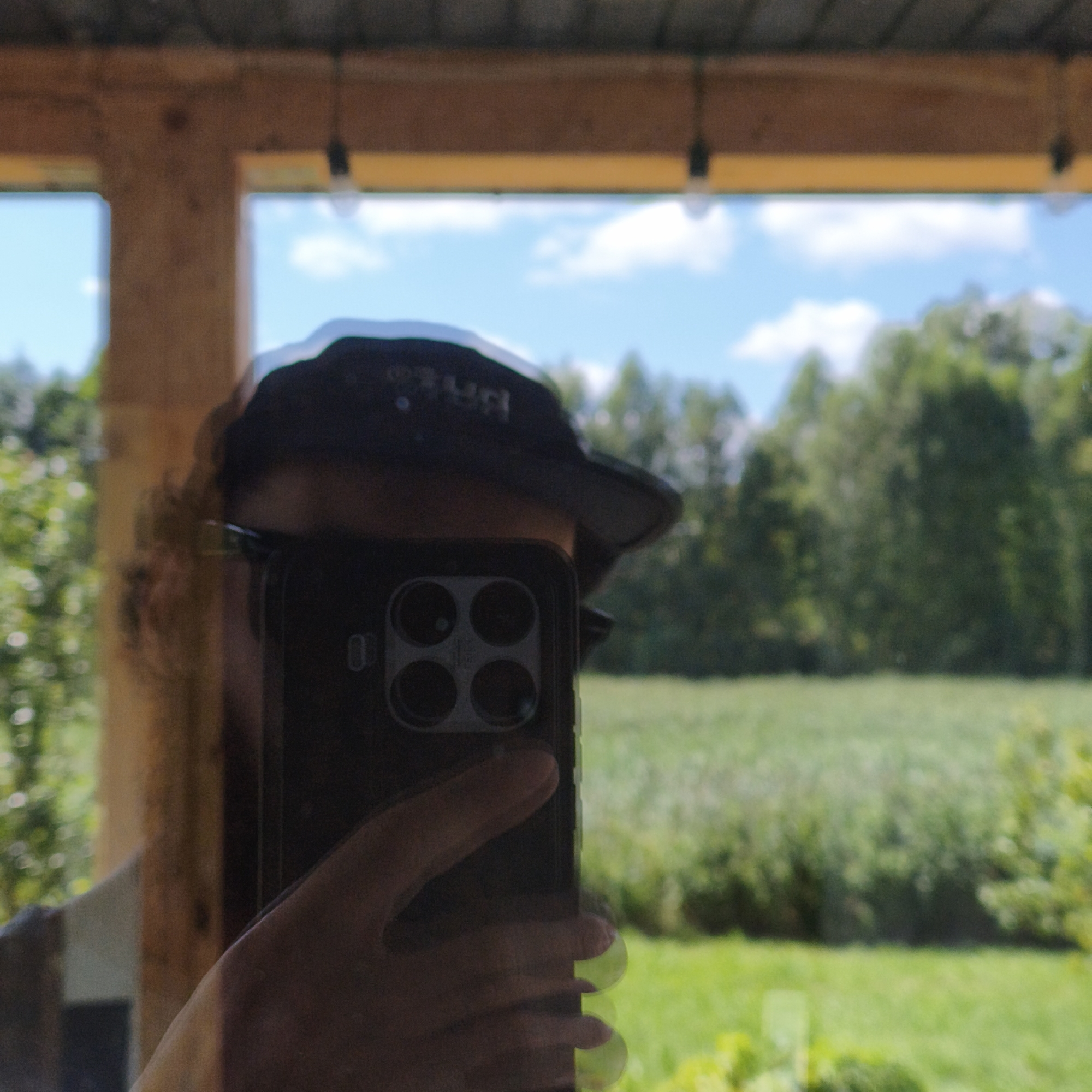The image is a reflection in a mirror showing a person holding a smartphone, taking a selfie. The person is wearing a dark baseball cap. The background visible in the reflection is an outdoor scene with green trees, a field, and a blue sky with clouds, suggesting a rural or suburban environment. There are wooden beams and a yellow trim visible at the top of the frame, indicating the presence of a structure like a porch or gazebo. The location is identified as Парэчча, Belarus. The smartphone has a prominent camera module with multiple lenses. The lighting suggests it is daytime.