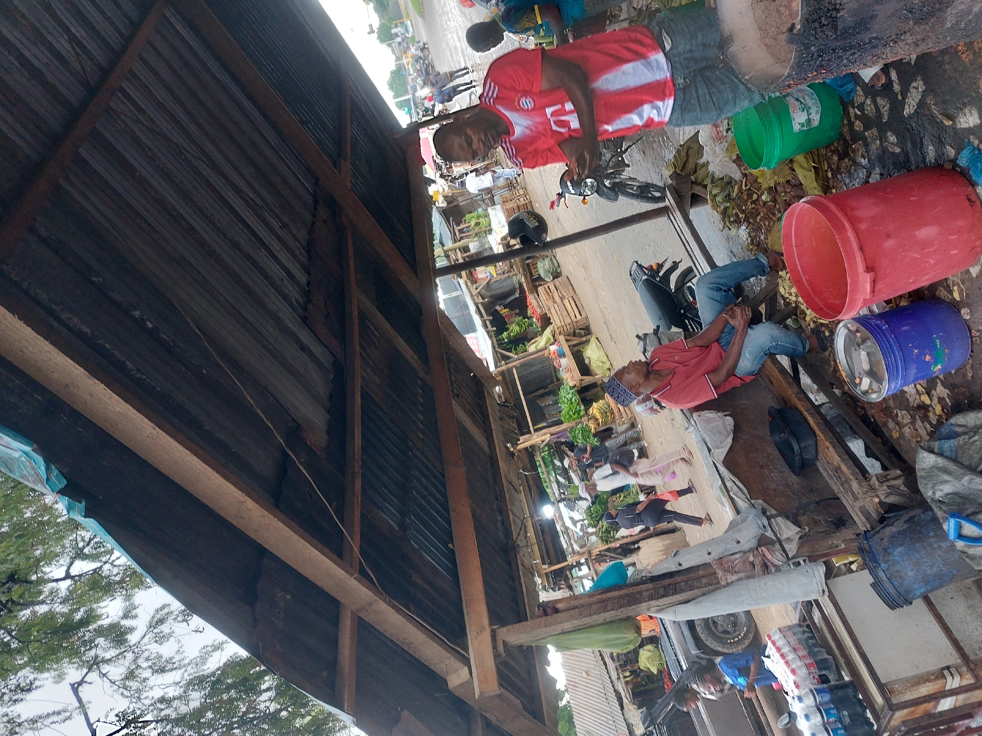 The image shows an outdoor market scene, seemingly in the city of Dar es-Salaam, Tanzania. The view is partially obscured by the corrugated iron roof of a market stall. Several people are present, engaged in activities related to selling and purchasing goods. Two men are prominently visible near the foreground: one wearing a red jersey is standing next to a motorbike and the other man is wearing a red shirt and seated on a structure. Nearby are plastic buckets of red, green, and blue colors. Other people are visible further in the background amongst rows of fruit and vegetable vendors. The environment appears to be a typical bustling marketplace.