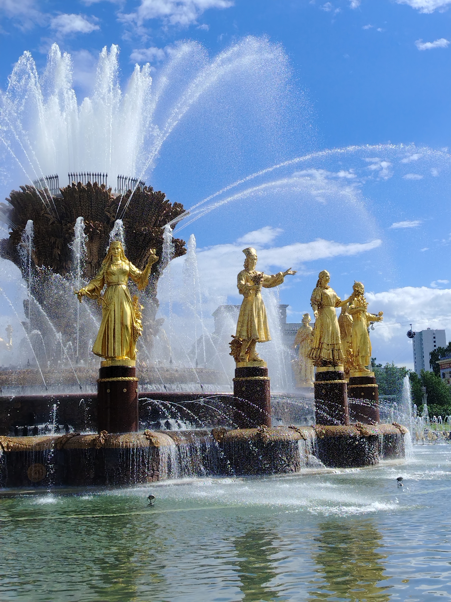 This media file features a majestic, golden fountain set against a vibrant blue sky with scattered white clouds, suggesting a bright, sunny day. The central subject is a large, ornate fountain with multiple tiers and dramatic water displays. Dominating the foreground are several gilded statues of women, each depicted in flowing robes and holding various symbolic items, such as grapes, flowers, or cornucopias. These statues stand on tall, cylindrical pedestals adorned with decorative elements.
The fountain itself is a spectacle of water, with powerful jets shooting high into the air, creating a mist that catches the sunlight. Water cascades down the tiered structure, creating a sense of movement and life. In the background, glimpses of modern buildings and lush green trees can be seen, providing context for an urban park or public square. The overall mood is one of grandeur and festivity. The image was likely captured in the afternoon due to the bright, direct sunlight and the clear sky. The style of the statues and the fountain design are indicative of Soviet-era monumental art, placing the likely location as Moscow, Russia, specifically at the VDNKh (Exhibition of Achievements of National Economy) park, which is famous for its "Friendship of Peoples" fountain. There is no visible text in the image.