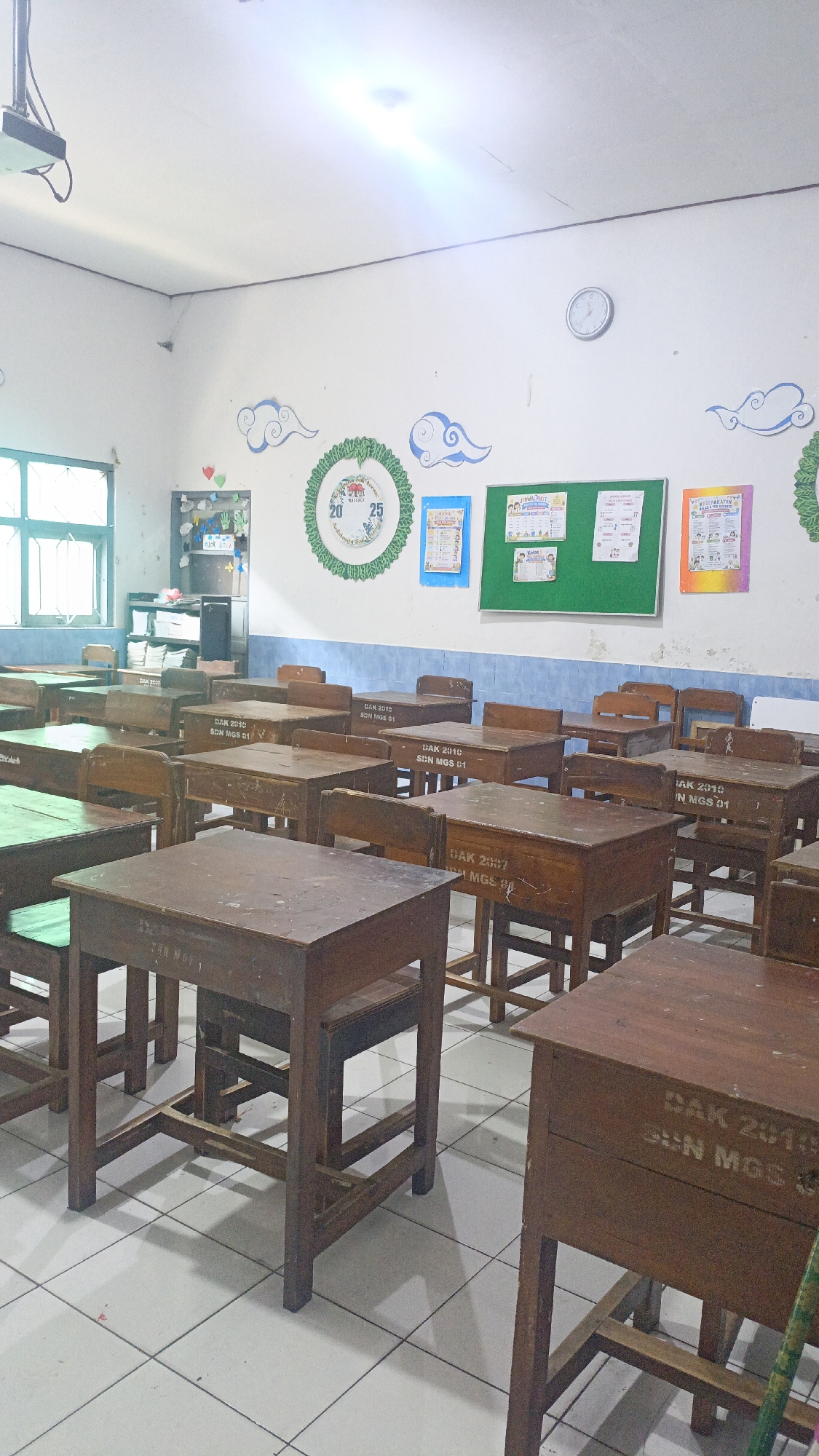 The image depicts an empty classroom in Salatiga, Indonesia. The room is filled with rows of wooden desks and chairs. The walls are white, with some decorations including a clock, a circular artwork with numbers 20 and 25, and several blue cloud cutouts. A green bulletin board is mounted on the wall, displaying various papers and notices. A projector is mounted on the ceiling. Natural light streams in from a window on the left side of the room, illuminating the tiled floor. The desks appear to be old and worn, with some bearing inscriptions. No people are visible in the scene.