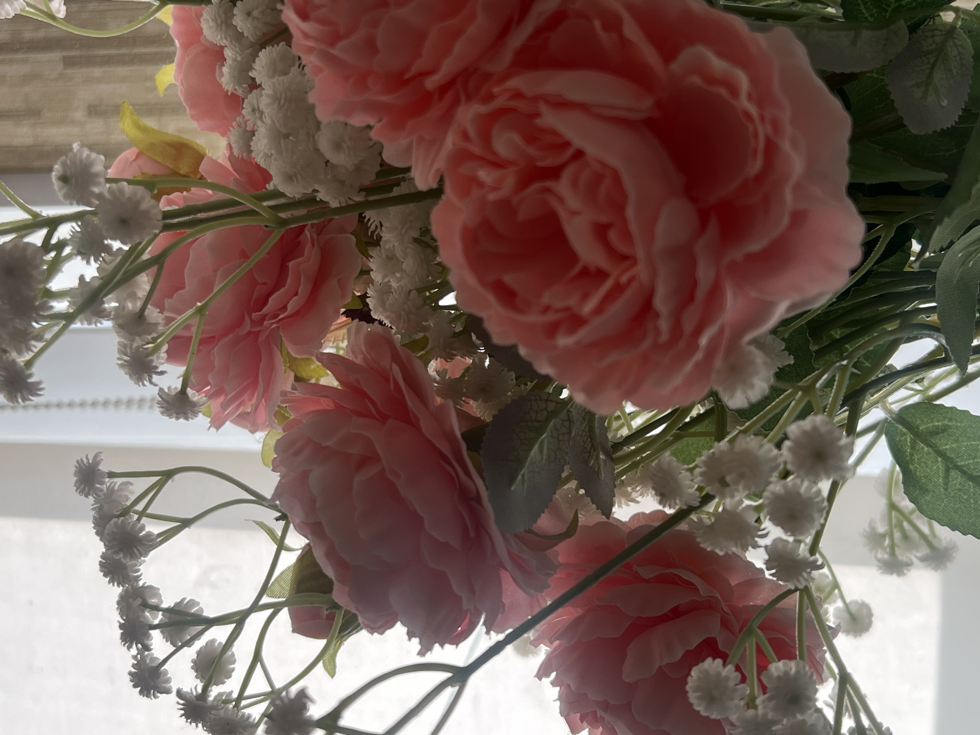 A close-up, slightly overexposed indoor shot shows a bouquet of artificial pink roses and white baby's breath flowers. The flowers are arranged in a vase, though the vase is not visible. The background is blurred and consists of a white curtain or blind on the left, and a textured, beige curtain or blind on the right. The lighting suggests the flowers are near a window. There are no people or animals present in the image. The scene depicts a decorative arrangement of flowers in a domestic or indoor setting. The location is Chaikovsky, Russia.