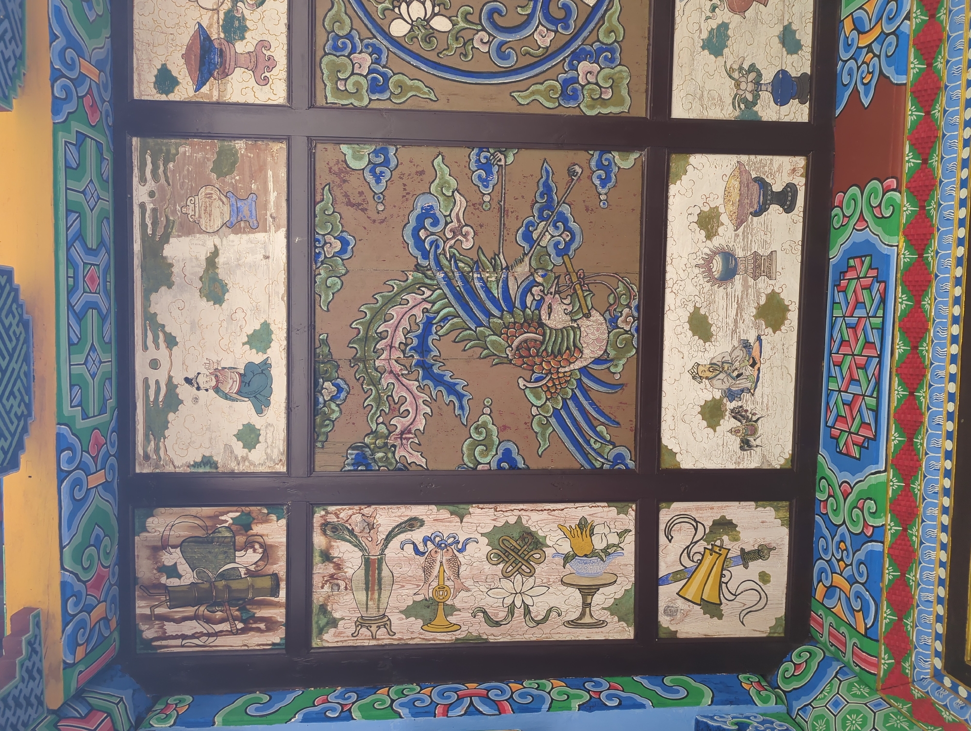 This image depicts the painted ceiling of a traditional building in Gucheng District, China. The ceiling is divided into multiple rectangular panels, each featuring intricate and colorful artwork.
The central panel displays a vibrant depiction of a mythical bird, possibly a phoenix, with blue and pink plumage, surrounded by swirling clouds. Other panels showcase various motifs:
* **Top row, left:** A figure in green robes is depicted in profile, standing next to what appears to be a wooden structure.
* **Top row, middle left:** A seated figure in blue robes holds an object in their hands.
* **Top row, middle right:** A blue and silver urn or vase sits on a pedestal.
* **Top row, right:** A red and blue conical object with intricate carvings rests on a pink pedestal, resembling a lamp or incense burner.
* **Middle row, left:** This panel contains a collection of objects, including a vase with peacock feathers, a fish with a coin emblem, a white flower, and a yellow flower on a pedestal.
* **Middle row, right:** A circular motif with a white flower and swirling blue and green patterns is visible.
* **Bottom row, left:** A bamboo stalk with a green leaf-like covering is depicted.
* **Bottom row, middle left:** A golden horn-like object and a sword are shown.
* **Bottom row, middle right:** A seated figure in traditional attire is depicted on horseback, with another seated figure next to them.
* **Bottom row, middle:** A blue urn or vase on a pedestal is visible, along with a patterned bowl.
* **Bottom row, right:** A blue vase holds a white flower.
The artwork exhibits a combination of detailed brushwork and stylized designs, characteristic of traditional Chinese art. The panels are separated by dark brown wooden frames. The surrounding architectural elements are also decorated with colorful geometric patterns and swirling motifs in blue, green, red, and yellow. The overall impression is one of rich cultural heritage and artistic craftsmanship.