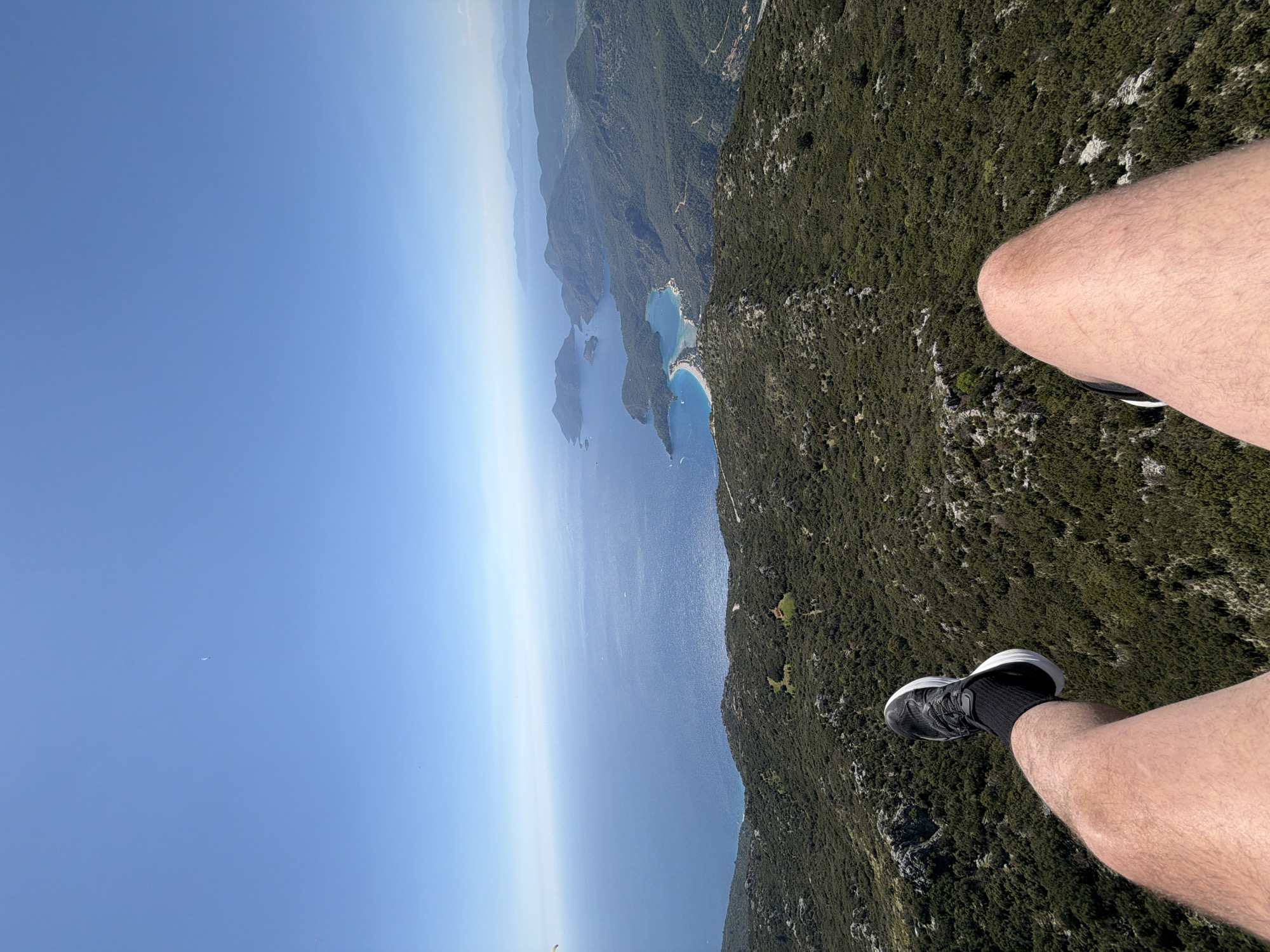 The image depicts a first-person perspective of someone paragliding, with their legs and feet visible in the foreground. They are wearing black athletic shoes with black socks. Below, a lush green landscape transitions into a coastline with multiple islands and bays featuring light-colored beaches. The ocean stretches into the horizon, meeting a clear blue sky. The location appears to be near Fethiye, Turkey, given the characteristic coastal scenery.
