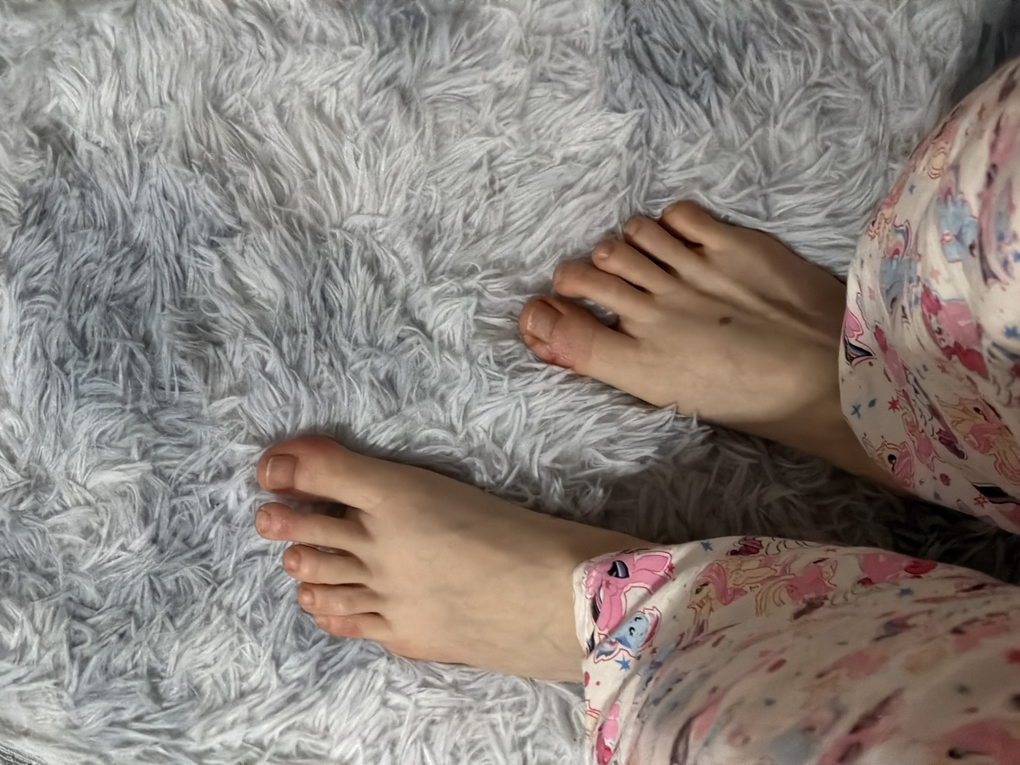 A close-up, overhead view shows a pair of bare feet resting on a plush, gray carpet. The feet are pale with visible toenails. The left foot is positioned slightly higher and closer to the viewer, with toes slightly curled. The right foot is positioned further down and to the right, with its toes more spread out. The individual is wearing pajama bottoms with a pastel pattern featuring cartoon unicorns and stars. The lighting is soft, and the background is entirely filled by the textured carpet. There is no information to suggest a location in Moscow, Russia.