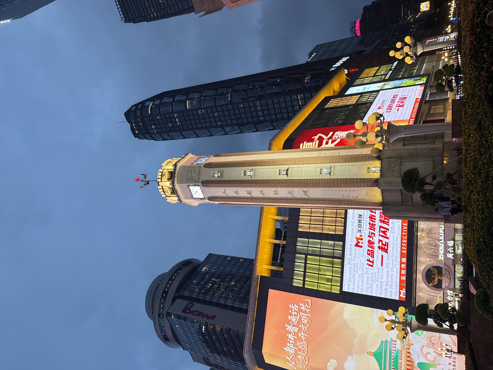 The image depicts a street scene at dusk in Yuzhong District, China, focusing on a clock tower and surrounding buildings. The clock tower is a prominent structure, illuminated and featuring a weather vane on top. It is surrounded by modern high-rise buildings and large digital billboards displaying advertisements. One billboard features a prominent red and white advertisement with Chinese characters. Another shows a stylized green building with a red roof. Streetlights with multiple globes illuminate the foreground, casting a warm glow on the manicured bushes and flowerbeds. Two individuals in uniforms, possibly security guards, are visible standing near the base of the clock tower. The sky is a deep blue, transitioning into twilight, with some clouds present. The overall impression is an urban environment at night, with a blend of traditional and modern architectural elements and dynamic commercial displays.