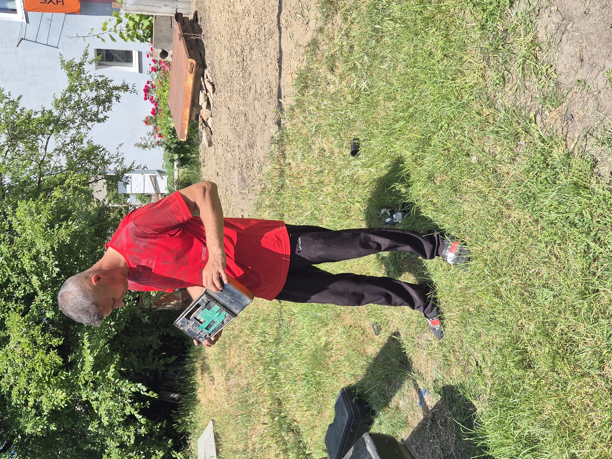 A man in a red t-shirt and black pants holds a disassembled piece of electronic equipment in a grassy outdoor area. He appears to be examining the components. In the background, a light blue house with white trim and a red flower bed are visible. The scene is set in the village of Sozonivka, Ukraine, on a sunny day.