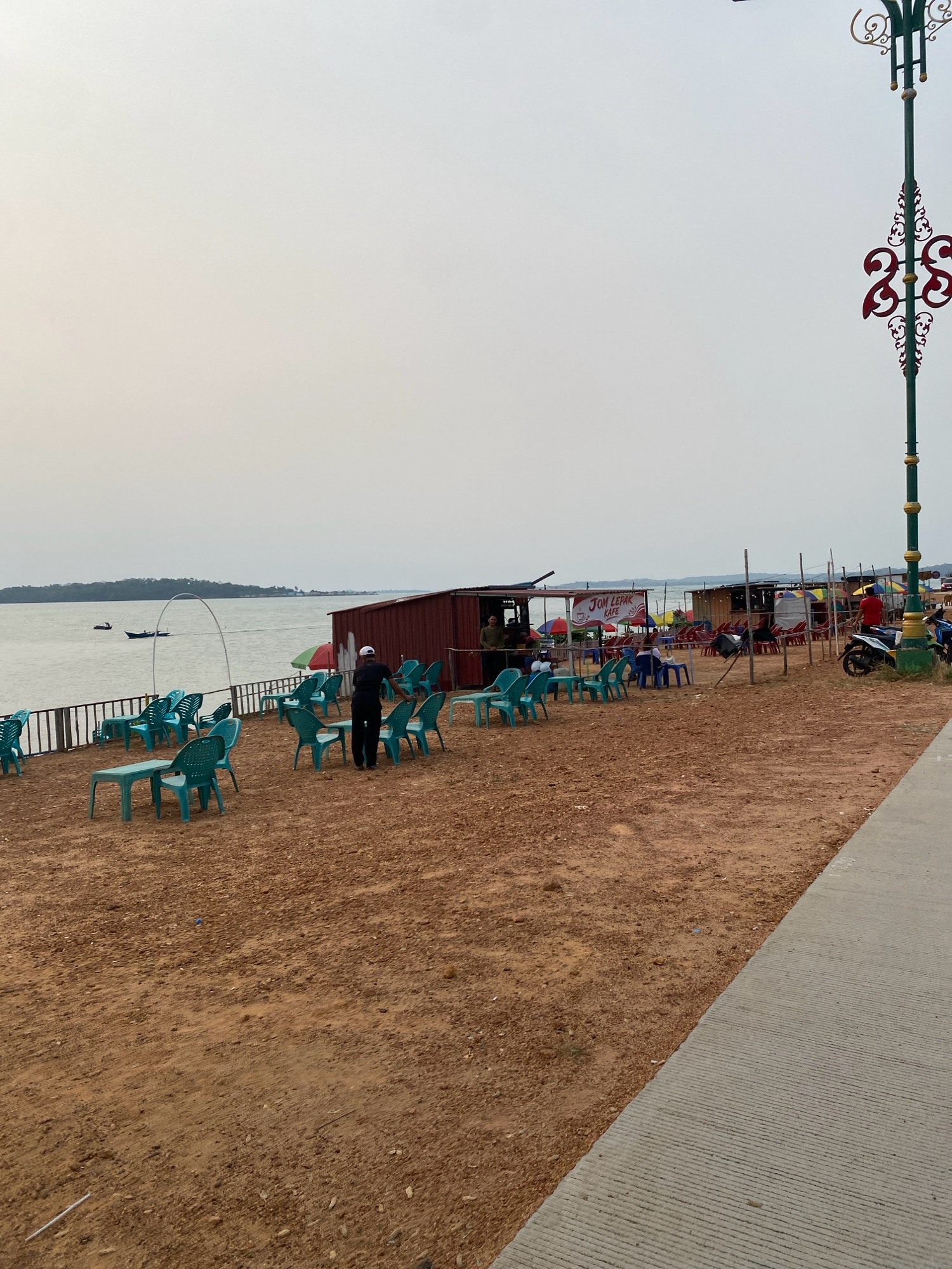 The image depicts a waterfront scene in Tanjung Pinang, Indonesia, likely during the late afternoon or early evening. The foreground is dominated by a sandy ground with several clusters of turquoise plastic chairs and tables, suggesting an outdoor seating area for a cafe or restaurant. A wooden fence runs along the edge of the sandy area, separating it from the water.
In the middle ground, a small, red-sided structure with a wooden roof appears to be a food stall or cafe, with a sign that reads "Jom Lepak Kafe." Several colorful umbrellas are visible, some providing shade over seating areas. People are present in the scene, including one individual standing near the red stall, and another person walking away from the camera near the water's edge.
The water itself is calm and reflects the hazy sky. A small boat is visible on the water in the distance, along with the outline of land on the horizon. To the far right of the image, a decorative lamppost with intricate metalwork stands tall, and a concrete walkway runs parallel to the sandy area. The overall atmosphere is one of a relaxed, open-air dining or gathering space by the water.