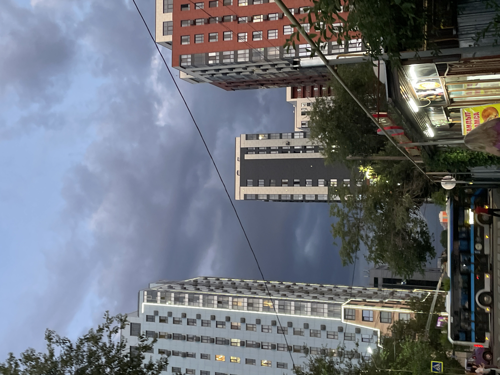 The image shows an urban scene with several multi-story buildings under a cloudy sky. The buildings are modern in design, with a mix of light blue, red, and grey facades, featuring numerous windows. Trees are scattered throughout the scene, some partially obscuring the buildings. A blue bus is visible on the right side of the frame, with its side facing the camera. A food stall or shop with bright lights and food items is also present. Some signs are visible, including one with Cyrillic text. The lighting suggests it is either dusk or a heavily overcast day. The scene appears to be a street in Almaty, Kazakhstan.