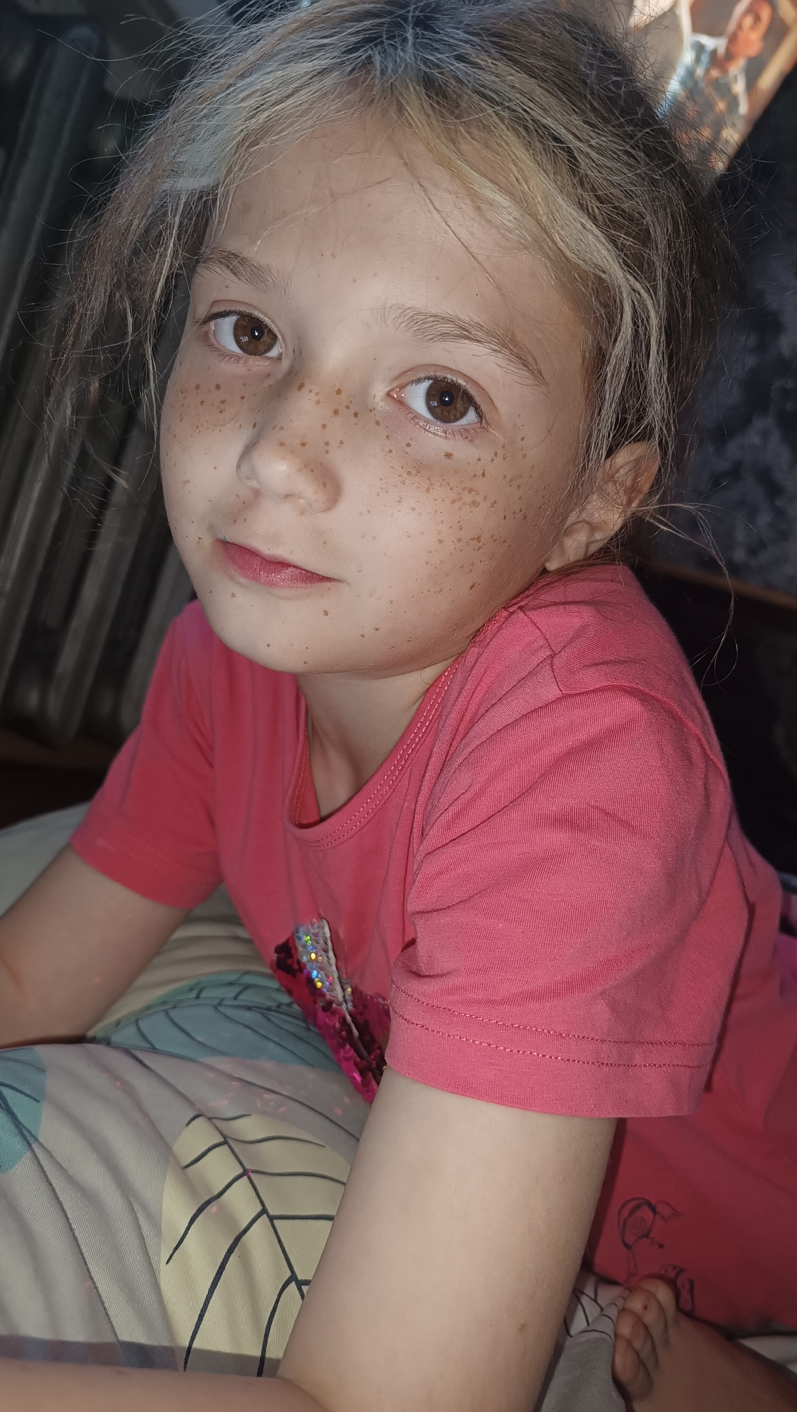 A medium shot shows a young girl with light brown hair and freckles looking towards the camera. She is wearing a pink t-shirt with a sequined design on the front. The girl is lying down on a patterned surface, possibly a bed or couch. Her left arm is resting on the surface, and her right arm is bent at the elbow, with her hand visible near her face. The background is dark and out of focus, with some vertical lines visible on the left, possibly a radiator or blinds. There is a framed picture visible in the upper right corner of the image, but the details are not clear. The image is a close-up, focusing on the girl's face and upper body. There is no information provided about the location in the city of Karsha, Russia.