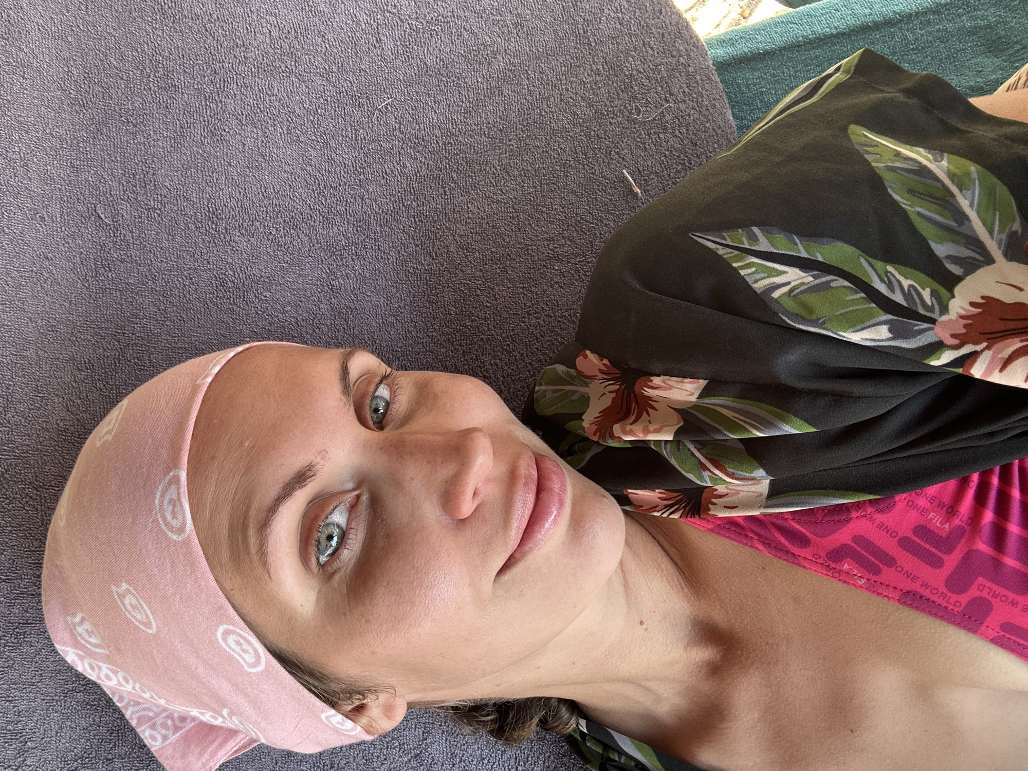 A fair-skinned woman with light blue eyes lies on a gray towel. She wears a light pink bandana and a floral-patterned top over a pink swimsuit top with the words "ONE WORLD ONE FILA" printed on it. The setting appears to be outdoors and bright. The precise location within Fethiye, Turkey, is not discernible from the image.