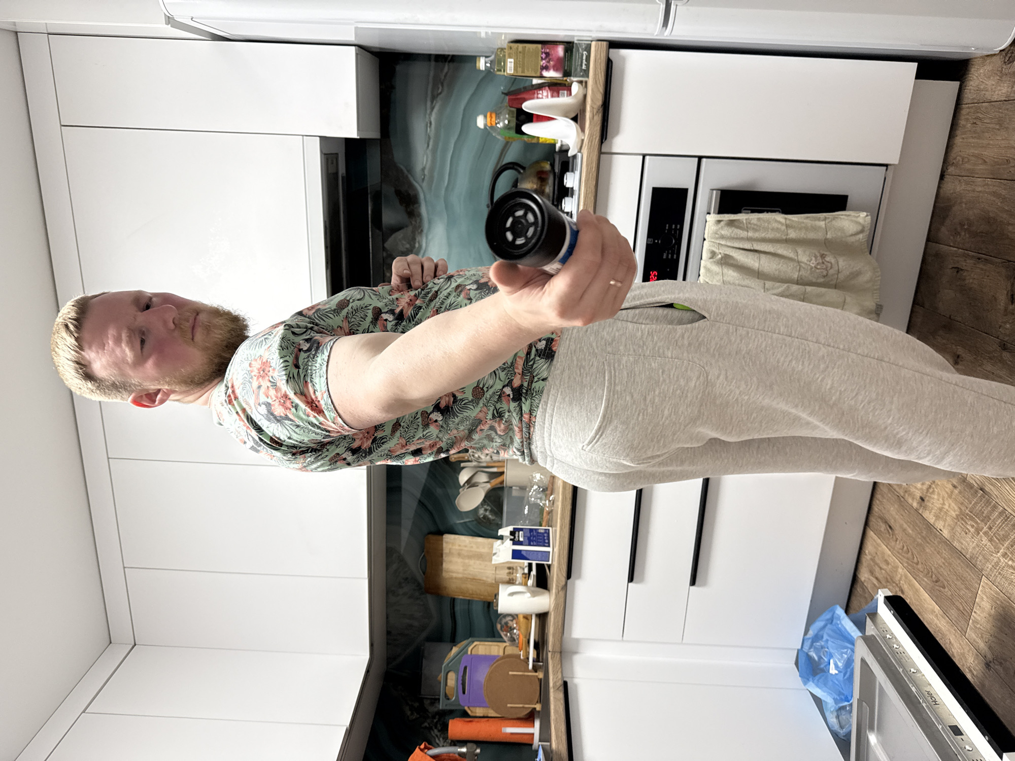 A fair-skinned adult male with blonde hair and a beard, wearing a short-sleeved t-shirt with a floral print and light grey sweatpants, is standing in a modern kitchen. He is holding a black cylindrical object with a silver ring and blue lettering in his right hand, extending it towards the camera. The man is looking over his left shoulder at the camera, with his body angled away.
The kitchen features white upper cabinets with no visible handles, and white lower cabinets with integrated black handles. The countertop is light wood. Behind the countertop, there is a backsplash with a teal, marbled pattern. Various kitchen items are visible on the countertop, including mugs, cutting boards, utensils, and bottles of cleaning supplies and oil. A white oven with a grey towel hanging from its handle is beneath the countertop. To the right of the oven, a white refrigerator is partially visible. The flooring is dark wood. In the lower left of the image, a blue trash bag is visible next to an open drawer or appliance, and a metallic tray is resting on the floor. The overall environment suggests a domestic setting. This image was likely taken in the city of Perm, Russia.