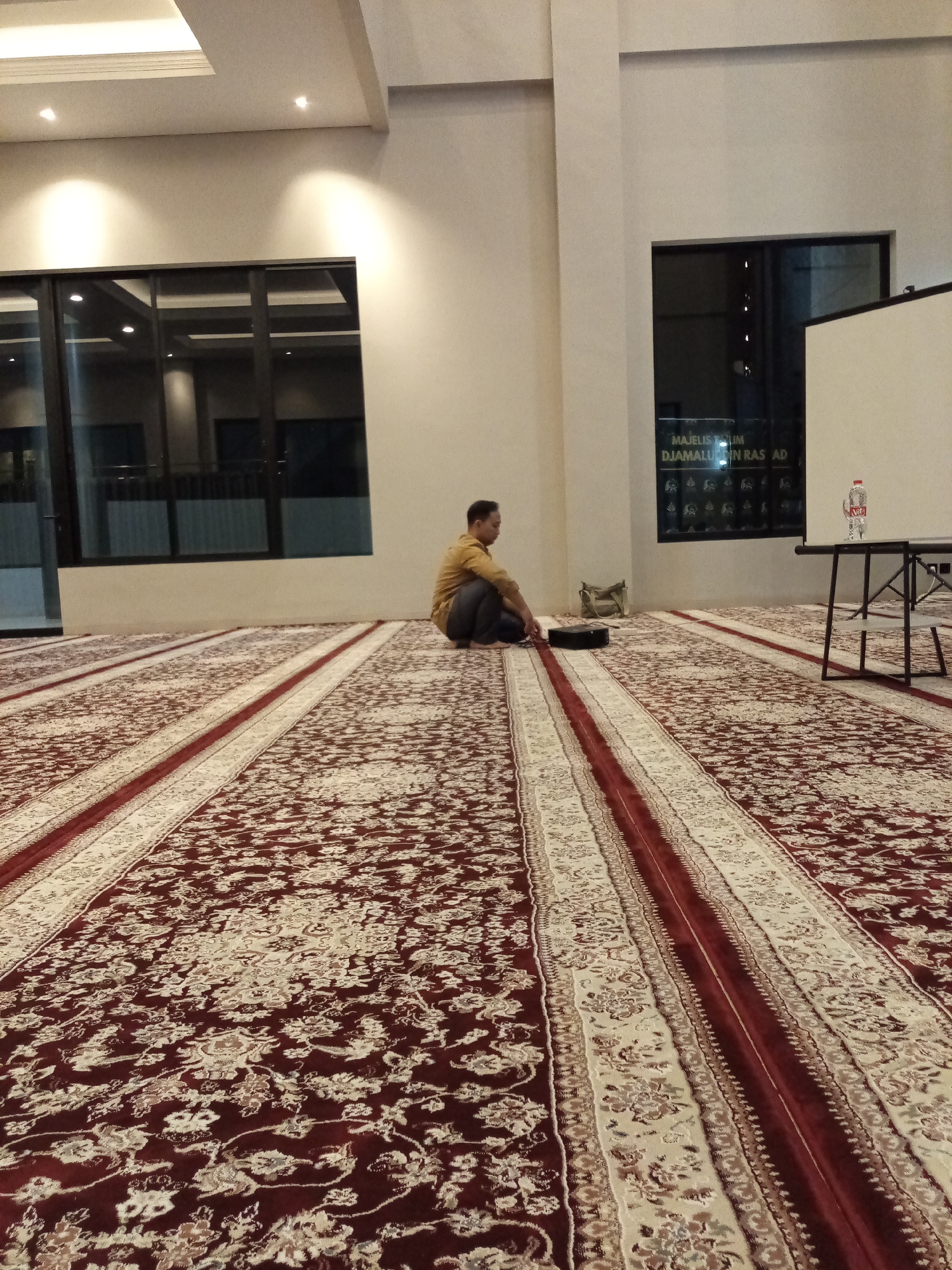 A man, appearing to be of Asian descent, wearing a mustard-colored long-sleeved shirt and dark pants, is sitting cross-legged on a large, ornate carpet within what appears to be a prayer hall or a large meeting room. The carpet features a complex floral pattern in shades of dark red, white, and beige, with prominent red and cream-colored striped borders running lengthwise. The man is positioned facing towards the left side of the image, with his hands resting on his lap. To his left, on the carpet, is a dark-colored electronic device, possibly a projector or speaker, and a beige bag.
The environment has large windows along the left wall, with dark frames and reflective glass showing interior lighting. The walls are a light, neutral color. In the background, there is a projector screen on the right side of the room, with a bottle of water on a dark table placed in front of it. A sign with text, partially visible and appearing to be in a foreign script, is mounted on the wall above and to the left of the screen. The ceiling has recessed lighting. The overall impression is of a well-lit indoor space with a formal carpet that dominates the foreground. The location is specified as Cihanjuang, Indonesia.