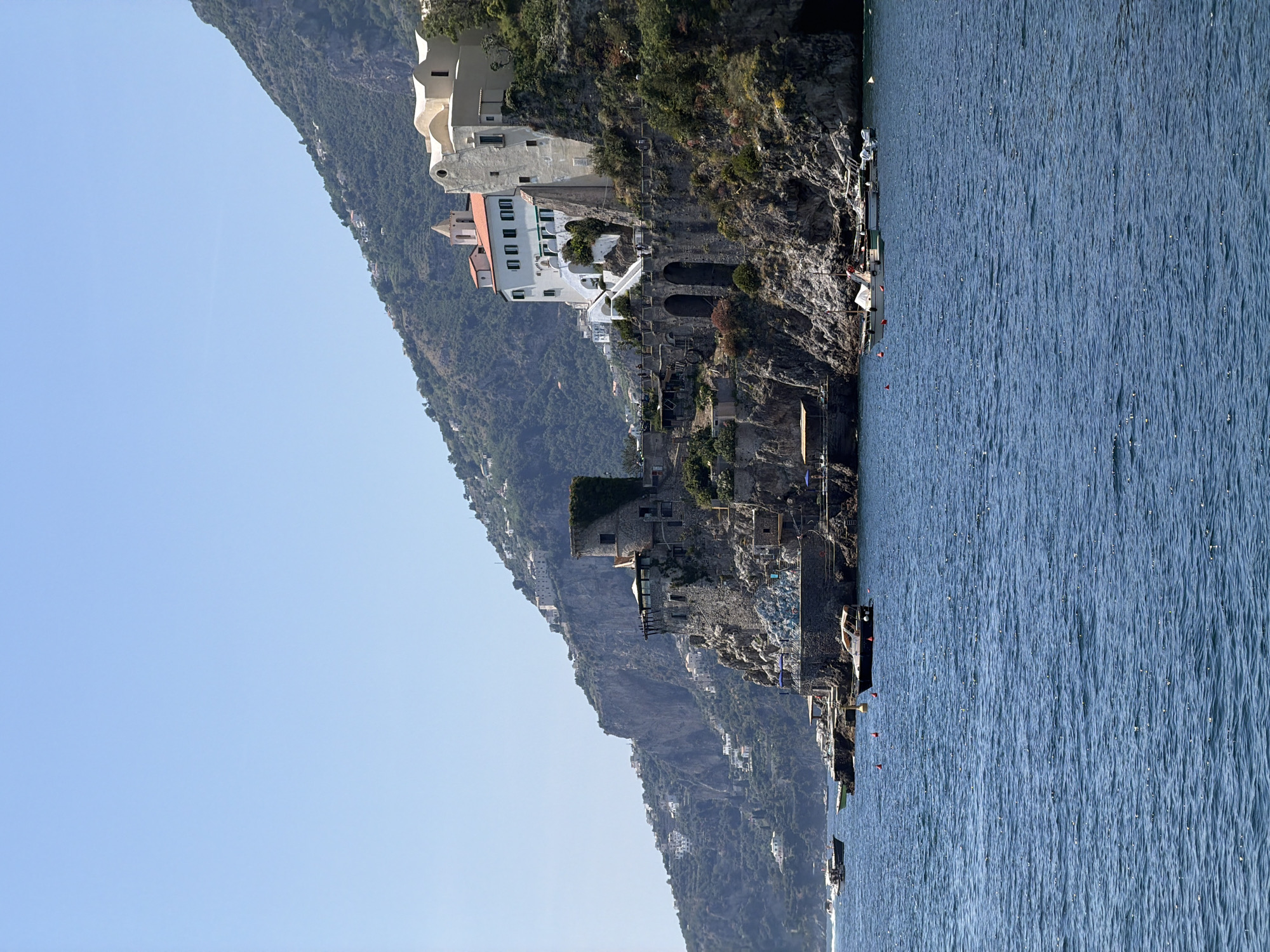 The image shows a coastal view in Ravello, Italy. A steep cliff face with built structures is the dominant feature. Buildings of various sizes, mostly light-colored, are built along the cliffside. The vegetation on the cliff is dense and green. Below the cliff, the blue water of the sea stretches into the distance. The sky is a clear, light blue, occupying a significant portion of the frame. There are boats docked along the rocky shoreline.