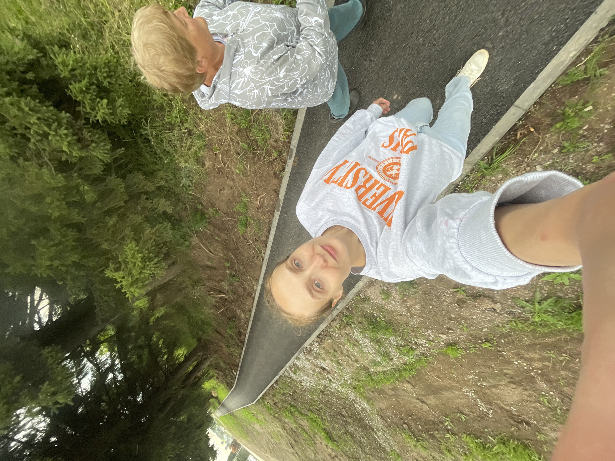 The media file shows two people from a high-angle perspective, walking on a paved path. The person in the foreground is looking directly at the camera, with a slight smile. They are wearing a light gray sweatshirt with orange text that reads "UNIVERSITY" in large, bold letters, and a pair of light blue jeans. The second person, further up the path and slightly to the left, is wearing a light gray sweatshirt with a white floral pattern and blue jeans. Both individuals appear to be outdoors. The surrounding environment consists of greenery, likely grass and trees, with the paved path running alongside a strip of dirt and grass. The lighting suggests it is daytime, and the weather appears to be overcast or subdued. The scene is captured from an unusual, tilted angle, adding a dynamic quality to the image. The location is identified as Sharkan, Russia.