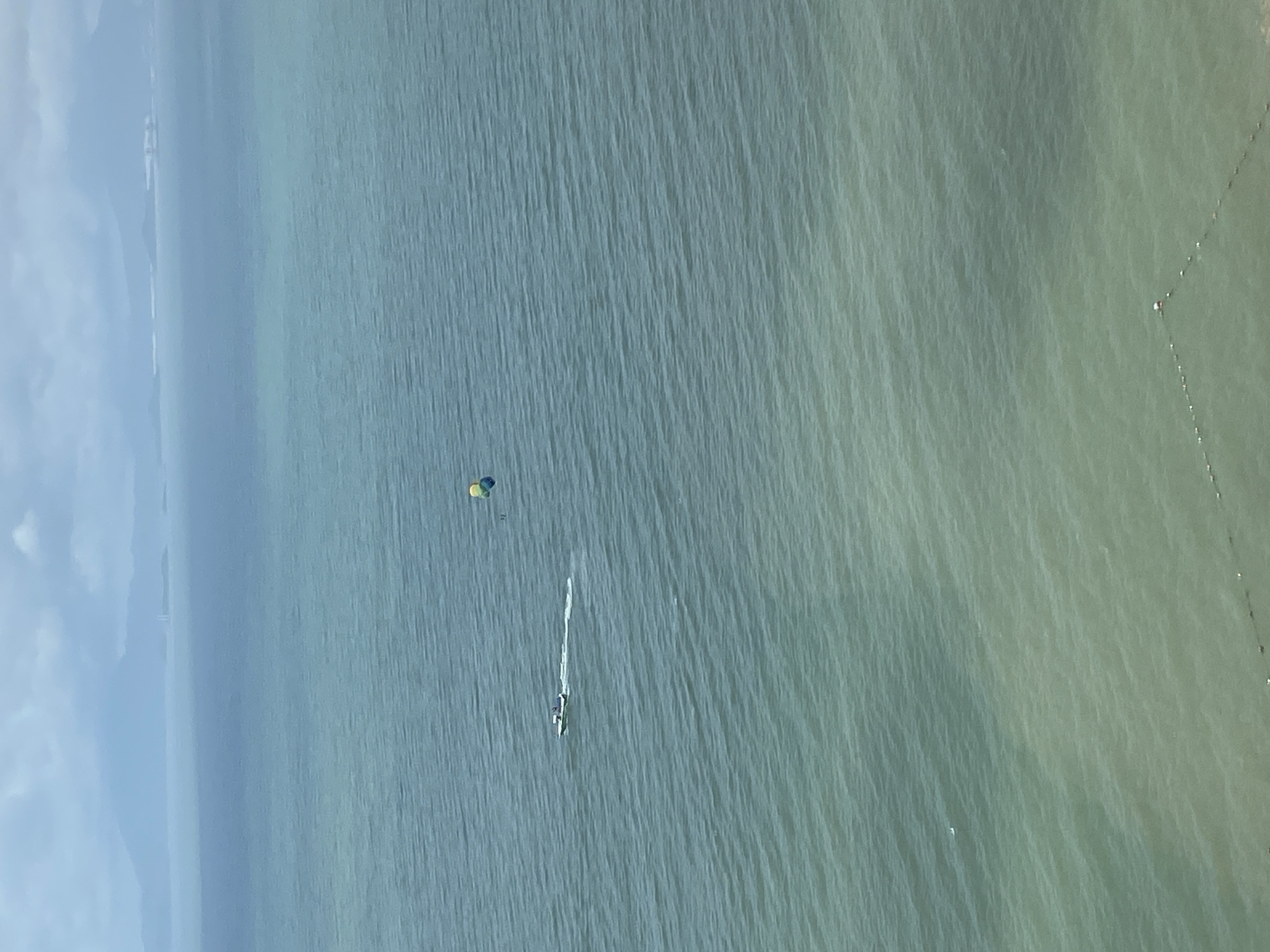 This image shows a wide expanse of ocean with a coastline in the distance, likely in Huizhou, China, given the presence of similar coastal scenery in the region. The water is a calm, blue-green color with gentle waves. In the mid-ground, a small white speedboat is moving, leaving a white wake behind it. Connected to the speedboat, a parasail with yellow, green, and blue sections is airborne. The parasail is positioned to the right of the boat. In the far distance, a hazy coastline with mountains and some structures is visible under a cloudy sky. The overall scene depicts a water sport activity on a partly cloudy day.