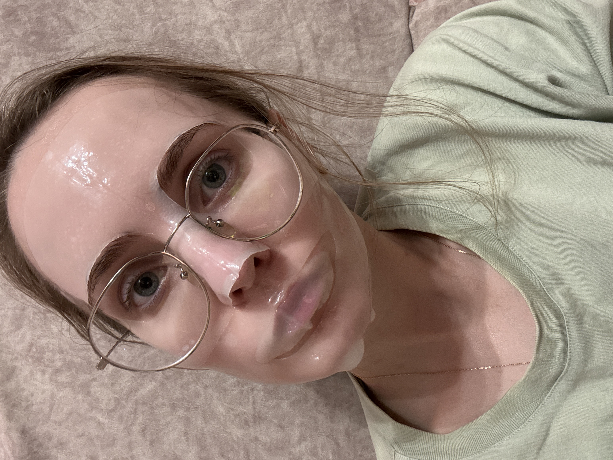 A young adult female is depicted in a close-up portrait, looking directly towards the viewer with a neutral expression. She has light-colored eyes, possibly blue or green, and light brown hair. Her face is covered with a translucent, light pink sheet mask, which appears to be saturated with serum and creates a glossy, moist surface. The mask has cut-outs for her eyes and nostrils, and partially covers her lips. Round, thin-framed metallic eyeglasses are worn over the face mask, resting on her nose and ears. She is wearing a light green, crew-neck top or t-shirt, and a delicate, thin gold-colored chain necklace is visible at her collarbone. The background consists of a soft, textured fabric surface in a light beige or pale pinkish-brown hue, likely upholstery. The lighting is soft and even. The image does not contain identifiable visual information to determine its location as Shirkovo, Russia.