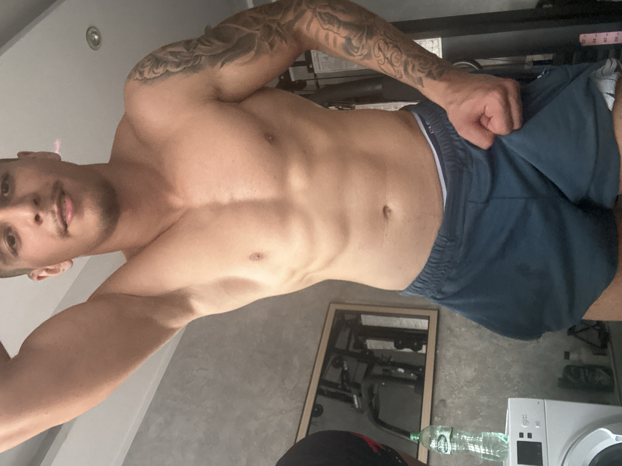 A man with dark hair and a tattoo on his left arm is holding up his blue shorts. The man is shirtless and has visible abs. In the background is a mirror with a reflection of workout equipment. A water bottle is sitting on top of a washing machine. The location is Unknown, Unknown.