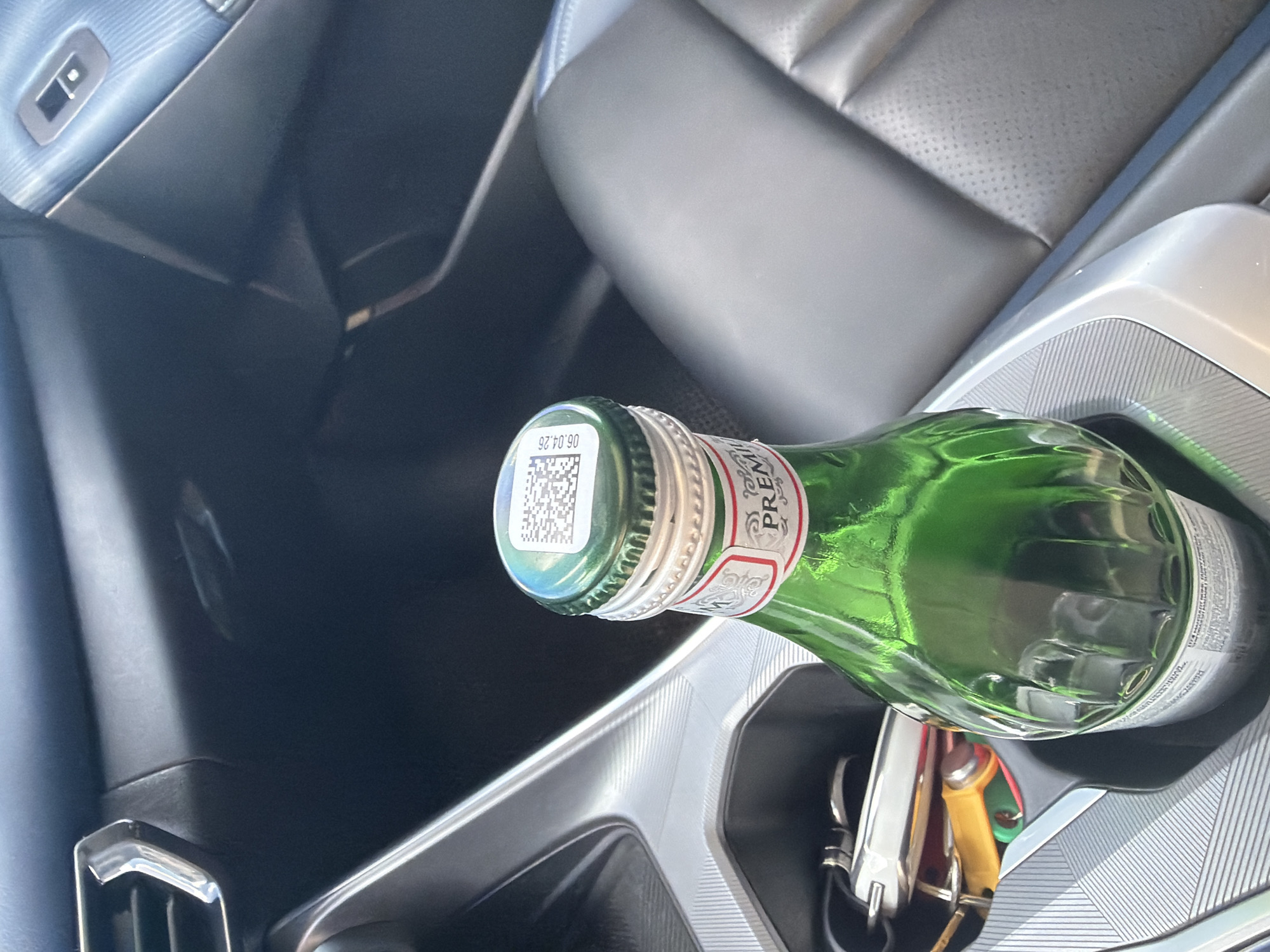 The image shows the interior of a car, with a green glass bottle of "Premium" brand sparkling water placed in a cup holder. The bottle has a silver screw-on cap with a QR code and the expiration date "06.04.26" printed on it. Keys are also visible in the cup holder next to the bottle. The car's dashboard and seats are visible in the background. The scene appears to be in the city of Chaikovsky, Russia.