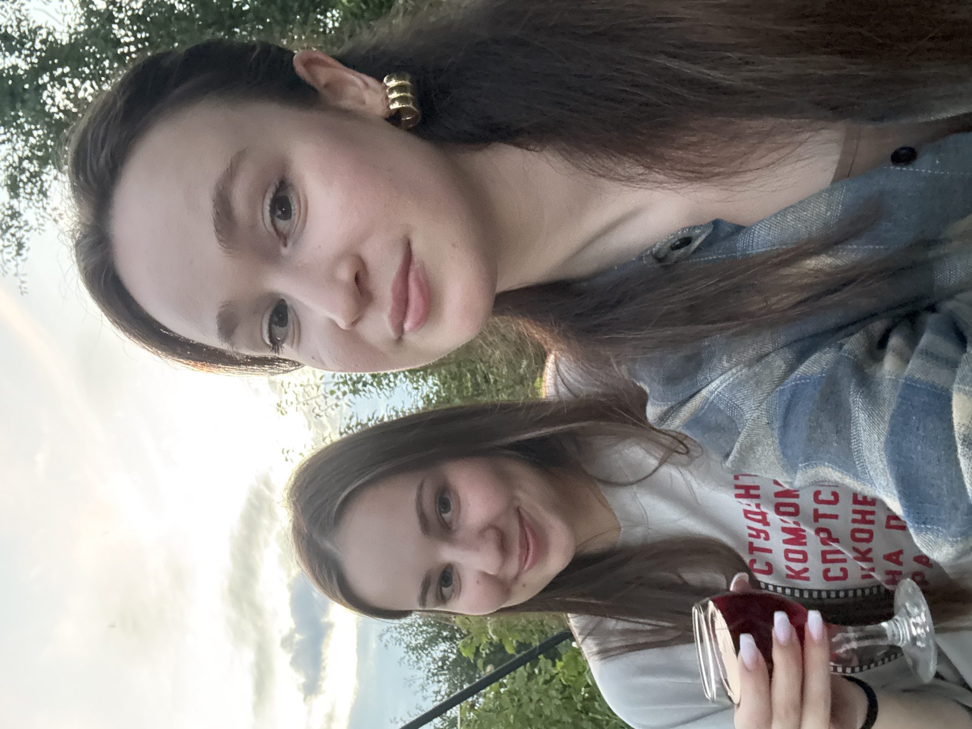 Two young women are taking a selfie outdoors in the city of Elovo, Russia, with trees and a cloudy sky in the background. The woman on the left is holding a stemmed glass of red wine. She is wearing a light-colored t-shirt with red text. The woman on the right is wearing a blue and grey plaid shirt over a tan top and has large gold hoop earrings. Both women have long brown hair and are smiling. The image appears to have been taken in the late afternoon or early evening, as indicated by the lighting.