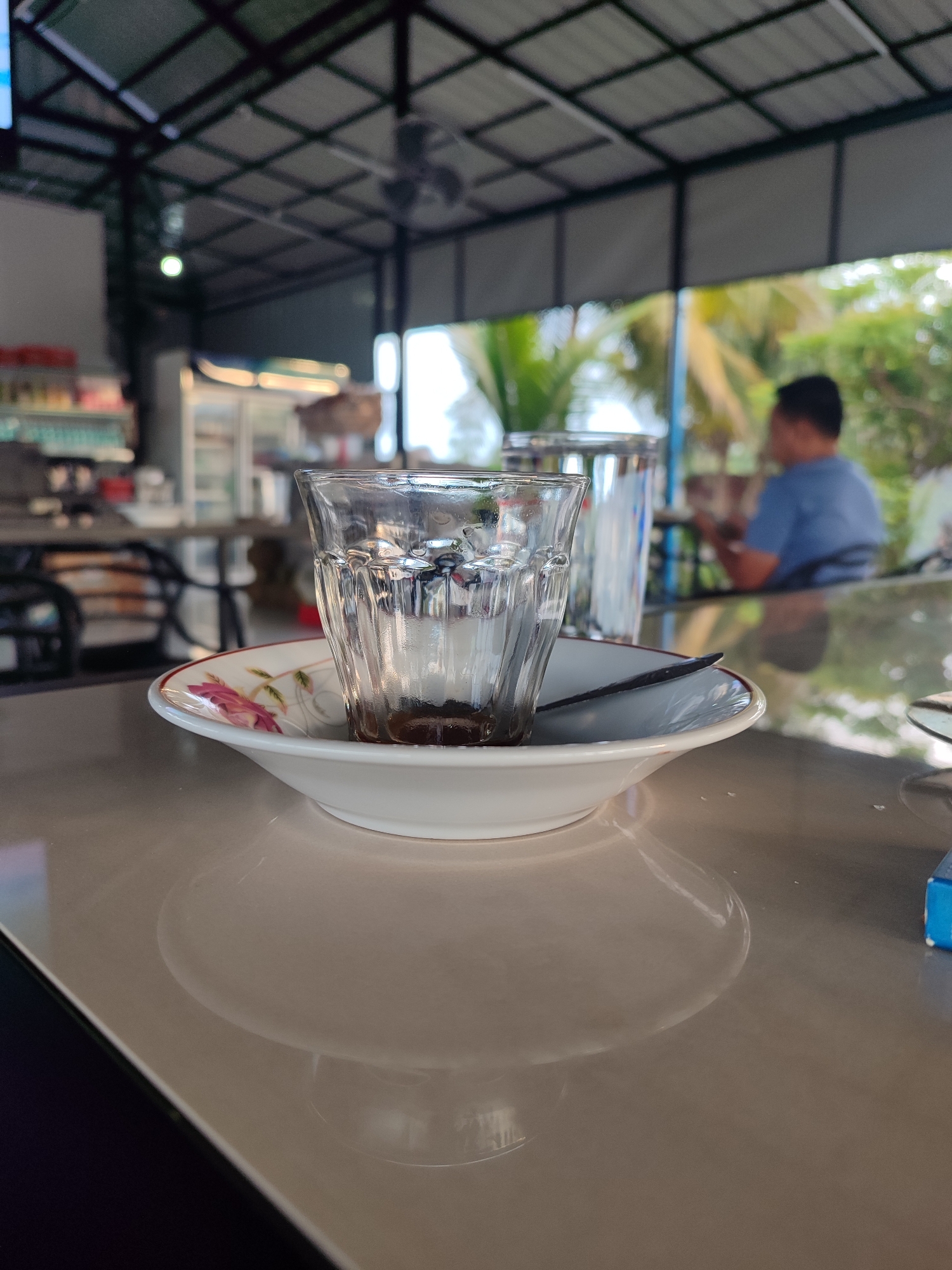 The image depicts a close-up, eye-level view of a small, partially filled glass of dark liquid, likely coffee, resting on a floral-patterned saucer. The glass is ribbed and shows condensation. A dark spoon is placed across the rim of the saucer. The setting appears to be an outdoor or semi-outdoor cafe or restaurant, with a corrugated metal roof supported by dark metal beams visible in the background. Beyond the roof, lush green foliage and palm trees suggest a tropical environment. In the blurred background, a person wearing a blue shirt is seated at a table, facing away from the camera. Other tables and chairs are visible, along with some shelves stocked with items, possibly in a cooler or display case, on the left side of the frame. The overall lighting is soft and diffused. The scene suggests a relaxed atmosphere in a place that appears to be located in a tropical region, possibly a coastal area. Based on the provided information, this scene is situated in Cotglumpang, Indonesia.