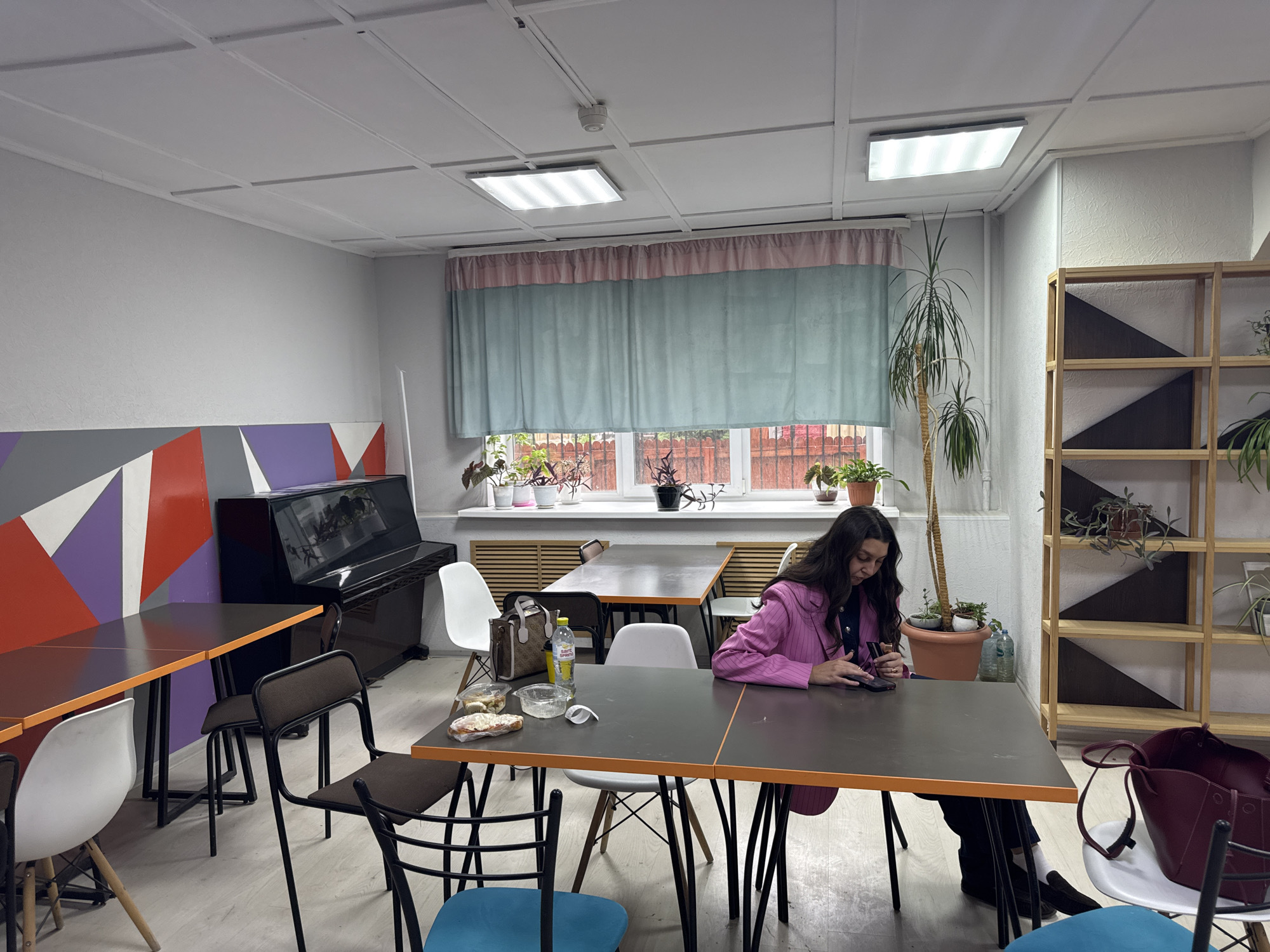 The image depicts the interior of a room, potentially a lounge or common area, in Perm, Russia. The location is suggested based on information about where the image was taken.
The room contains multiple tables with orange edges and black tops, each surrounded by chairs of various colors and designs, including white, brown, and blue. A woman wearing a pink blazer sits at one of the tables, focused on a smartphone in her hands. On the table, there are food containers, a bottle of water, and other miscellaneous items.
In the background, there's a black upright piano next to a wall with geometric patterns of orange, gray, and purple. A window adorned with a light blue curtain and a pink decorative valance is lined with potted plants. To the right of the window, a tall plant stands near a wooden shelf filled with additional plants. The room is lit by rectangular ceiling lights. The overall environment is somewhat cluttered but functional.