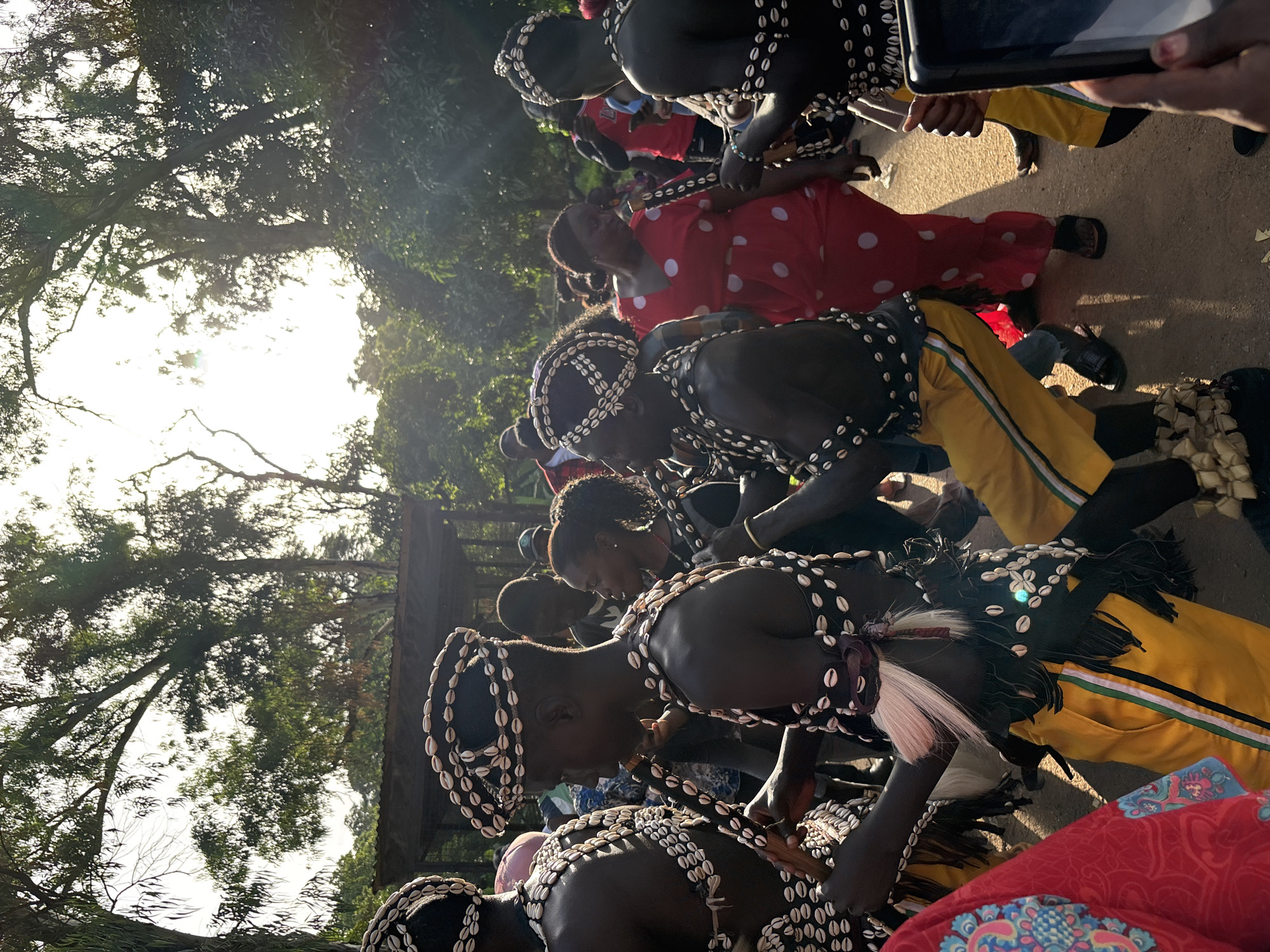The image shows a group of people in what appears to be a cultural or ceremonial setting in Jos, Nigeria. The subjects are adorned in traditional attire, featuring black fabric with intricate patterns of shells and other adornments, along with yellow trousers. Some individuals are wearing headdresses made of shells. The sun is bright, suggesting it is daytime, likely late morning or afternoon, with sunlight filtering through the trees, creating a dappled effect.
The people are positioned in a line or procession, with some looking down and others seemingly engaged in a ritual or performance. One person in the foreground is wearing a red and white polka dot dress. Another person is holding a tablet device, possibly capturing the event. The ground appears to be a dirt or gravel path. The surrounding environment includes lush green trees, indicating an outdoor setting. The overall mood seems to be one of solemnity or reverence associated with the cultural event. No visible text is present in the image.