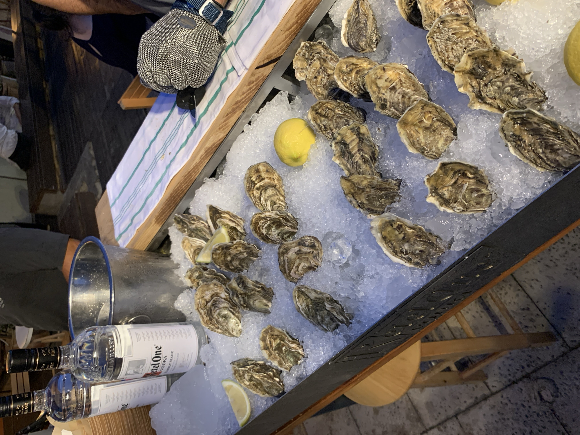 The image shows an assortment of fresh oysters arranged on a bed of crushed ice. A gloved hand holding an oyster shucking tool is present, suggesting someone is preparing the oysters. A stainless steel ice bucket and two bottles of Ketel One vodka are positioned nearby, indicating a pairing of oysters and vodka. A striped towel rests on the wooden counter. A half-cut lemon is nestled among the oysters. The wooden counter is likely part of an outdoor setup. While the location is stated as Tel Aviv, Israel, there are no distinct identifying features to verify this.