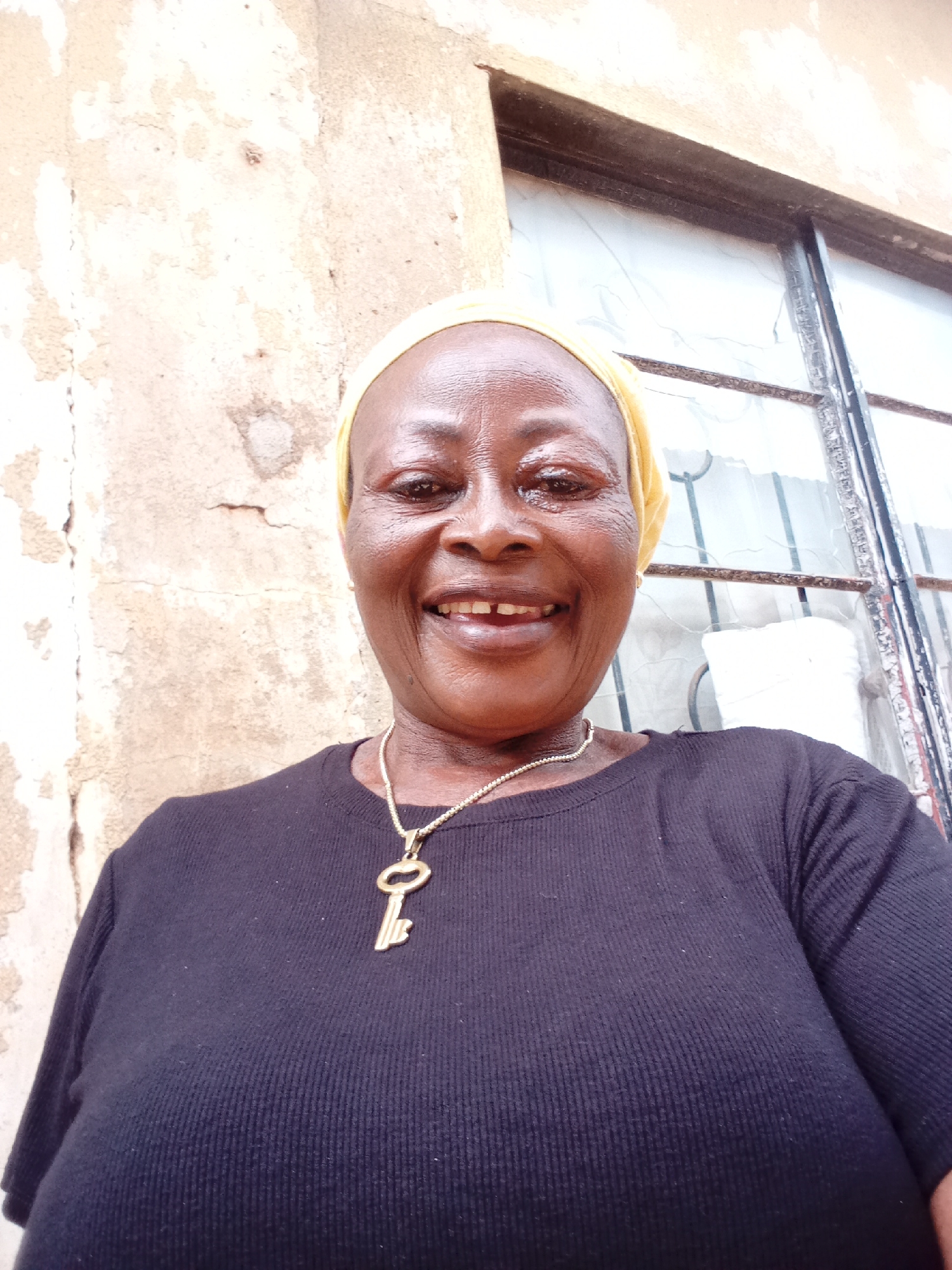 A medium shot selfie shows a Black woman with a yellow headscarf, a black t-shirt, and a gold key necklace. She has a broad smile with visible teeth and is looking directly at the camera. The background appears to be an outdoor setting with a textured, light-colored wall and a window with dark panes. The scene suggests a casual, everyday moment. The location is identified as Yelwa, Nigeria.