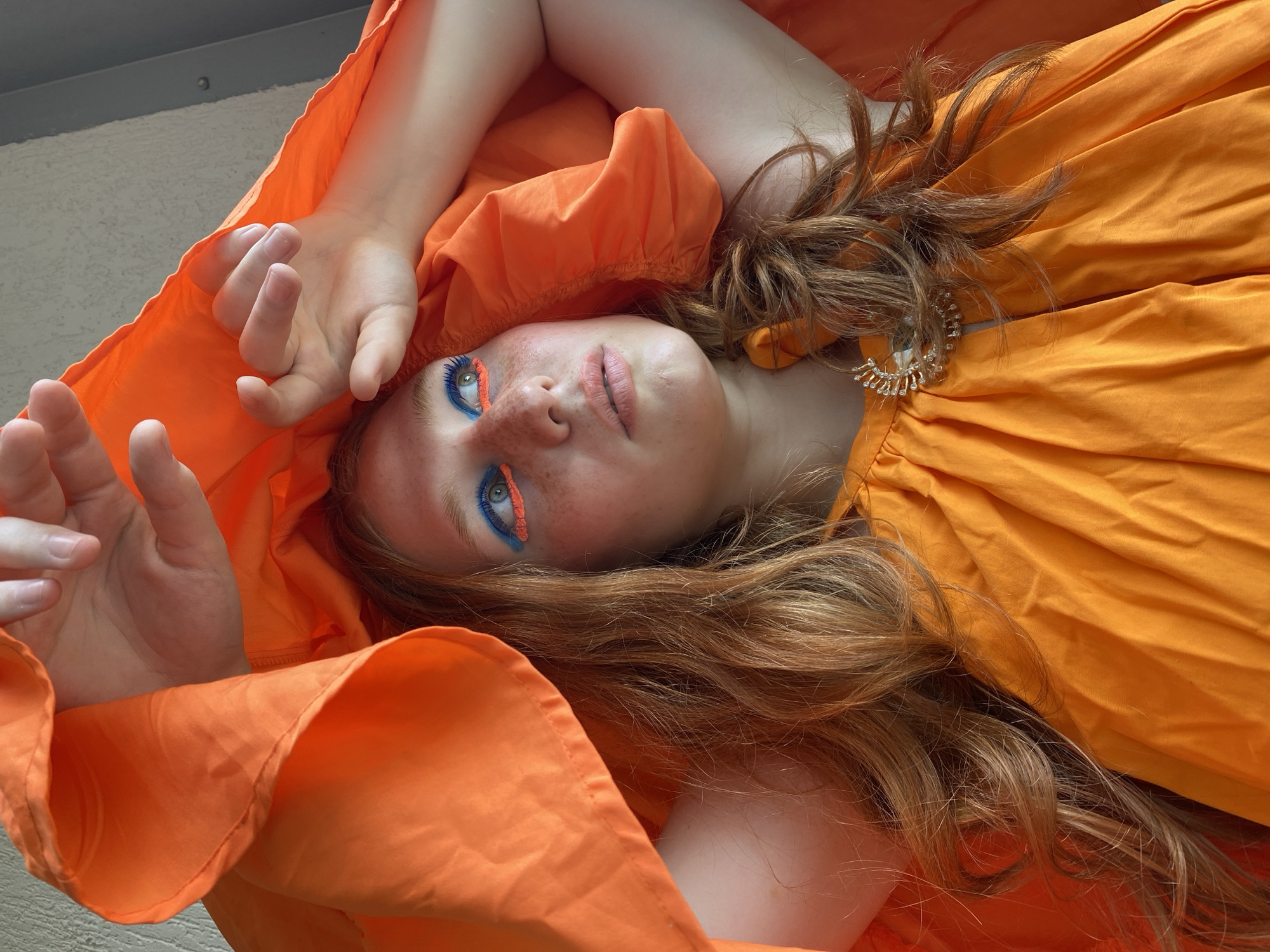 This media file features a young person with red hair, lying on their back, draped in a vibrant orange fabric that appears to be a dress or a large piece of clothing. The fabric is voluminous and gathered, creating a dramatic framing effect around the subject. The person is looking directly at the camera with a neutral expression, their eyes are adorned with striking blue and orange eye makeup. Freckles are visible on their nose and cheeks. Their hands are positioned near their face and head, one palm open and slightly raised, the other arm bent with the hand resting near their head. The background is out of focus but appears to be an outdoor setting with a textured wall and a grey, possibly metallic, surface above. The lighting suggests it is daytime, with soft shadows. The subject is wearing a sparkling brooch on the orange fabric near their shoulder. No visible text is present in the image. The overall aesthetic is artistic and expressive, with a focus on the striking makeup and the vivid color of the fabric.