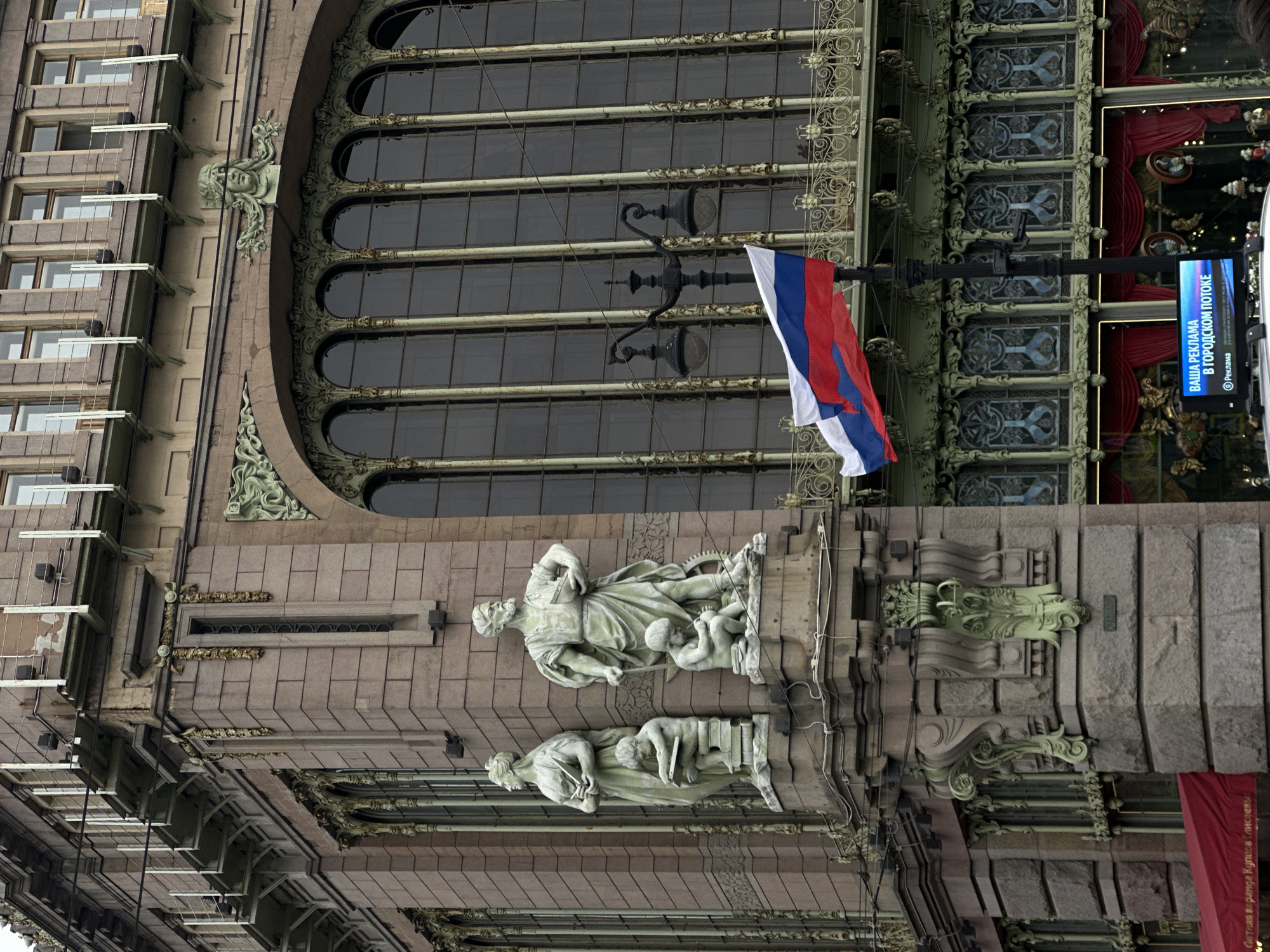 The image captures the Singer House, a historic building located at Nevsky Prospekt 28 in Saint Petersburg, Russia. The facade of the building is constructed from stone and features ornate architectural details, including large arched windows with intricate metalwork, decorative sculptures of figures and faces, and elaborate green metal embellishments. On the left side of the building, two stone sculptures of a woman and a man holding books stand on pedestals, flanked by smaller sculptures of children seated on stacks of books. A Russian flag is visible in the foreground, attached to a lamppost. Below the flag, a digital advertising screen displays text in Russian. The building's architecture suggests a late 19th or early 20th-century style. The sky is overcast, contributing to a subdued lighting condition.