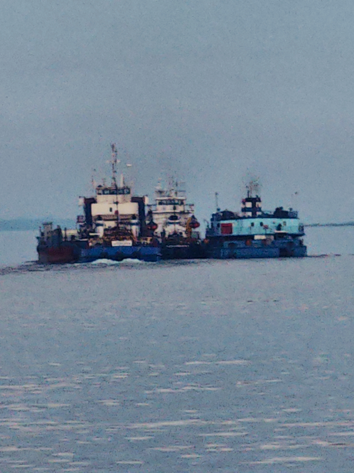 The image shows a calm body of water, likely a river or lake, with two tugboats or workboats moving side-by-side. The sky is overcast and a pale blue-gray, suggesting a cloudy day. The water is a muted blue-gray with some white reflections.
The tugboat on the left is larger and appears to be made of darker colored material, possibly red and dark blue, with a white superstructure. It has several decks and a mast with visible equipment. The tugboat on the right is smaller and brightly painted in turquoise and blue, with a red door on its side. It also has a superstructure with a mast and some visible equipment. Both vessels appear to be moving forward, creating a slight wake behind them.
The overall scene is serene and functional, depicting maritime activity. There are no people clearly visible on the boats. The location appears to be in Russia, given the provided context of Vorogovo. There is no visible text in the image. The time of day is difficult to determine due to the overcast sky, but it appears to be daytime. The weather is mild and uneventful.