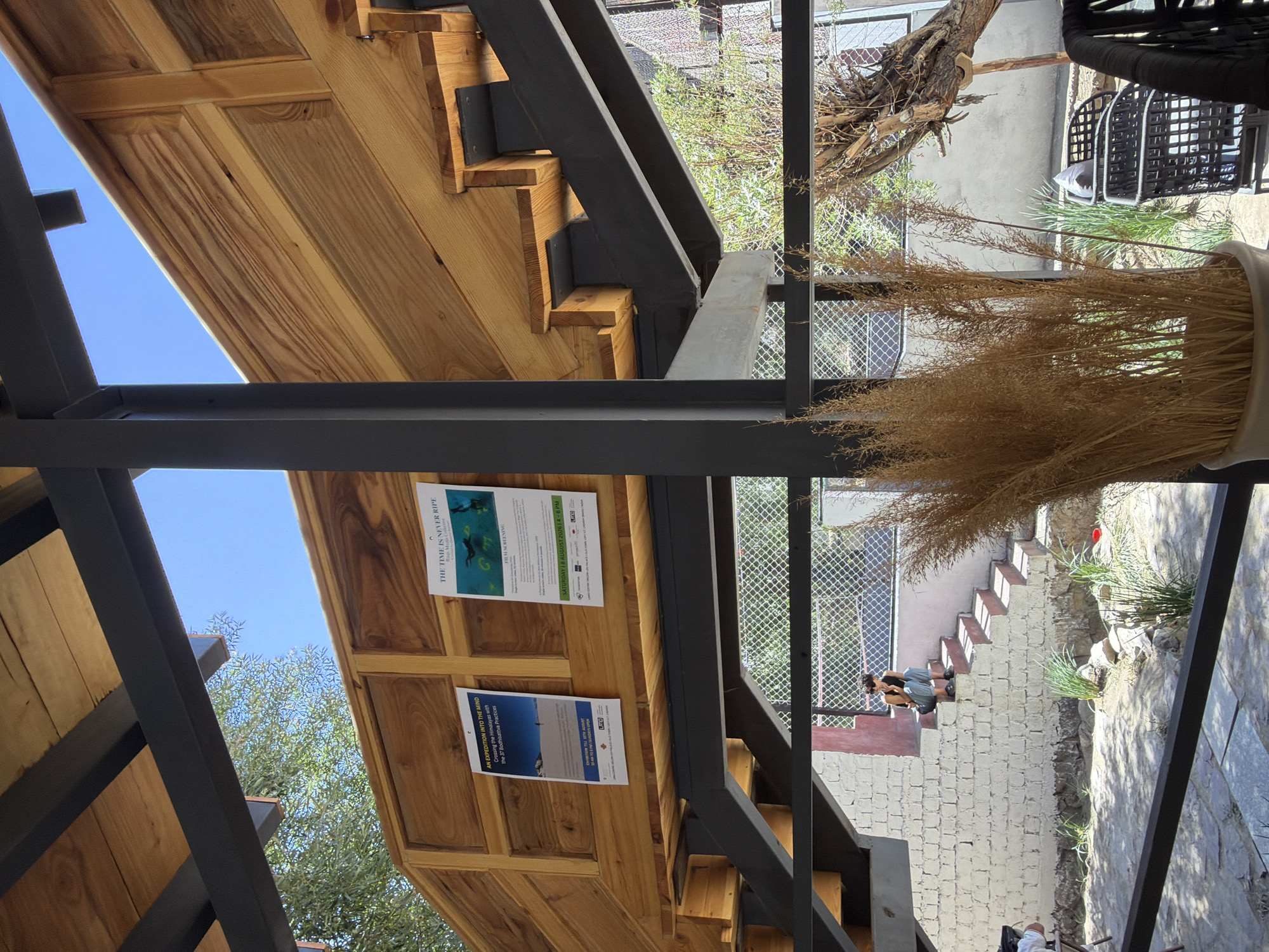 This image depicts an outdoor seating area in Leh, India, with a view of a multi-level wooden and metal structure that appears to be part of a building or an elevated walkway. Two posters are affixed to the wooden wall of this structure. The poster on the left, titled "AN EXPEDITION INTO THE MIND," details a crossing of the Himalayas with 37 Dobhachattva Practices. The poster on the right, titled "THE TIME IS NEVER RIPE," announces a film screening.
Below the elevated structure, a person is seated on a set of concrete steps. The steps lead down to a courtyard with a stone-paved ground, sparsely populated with green plants and some rocks. A large dried grass plant in a pot is in the foreground, partially obscuring chairs made of black woven material. The overall scene suggests a relaxed, open-air environment, possibly a cafe or a cultural space in Leh, a city located in the Indian Himalayas known for its Tibetan Buddhist heritage and stunning landscapes.
