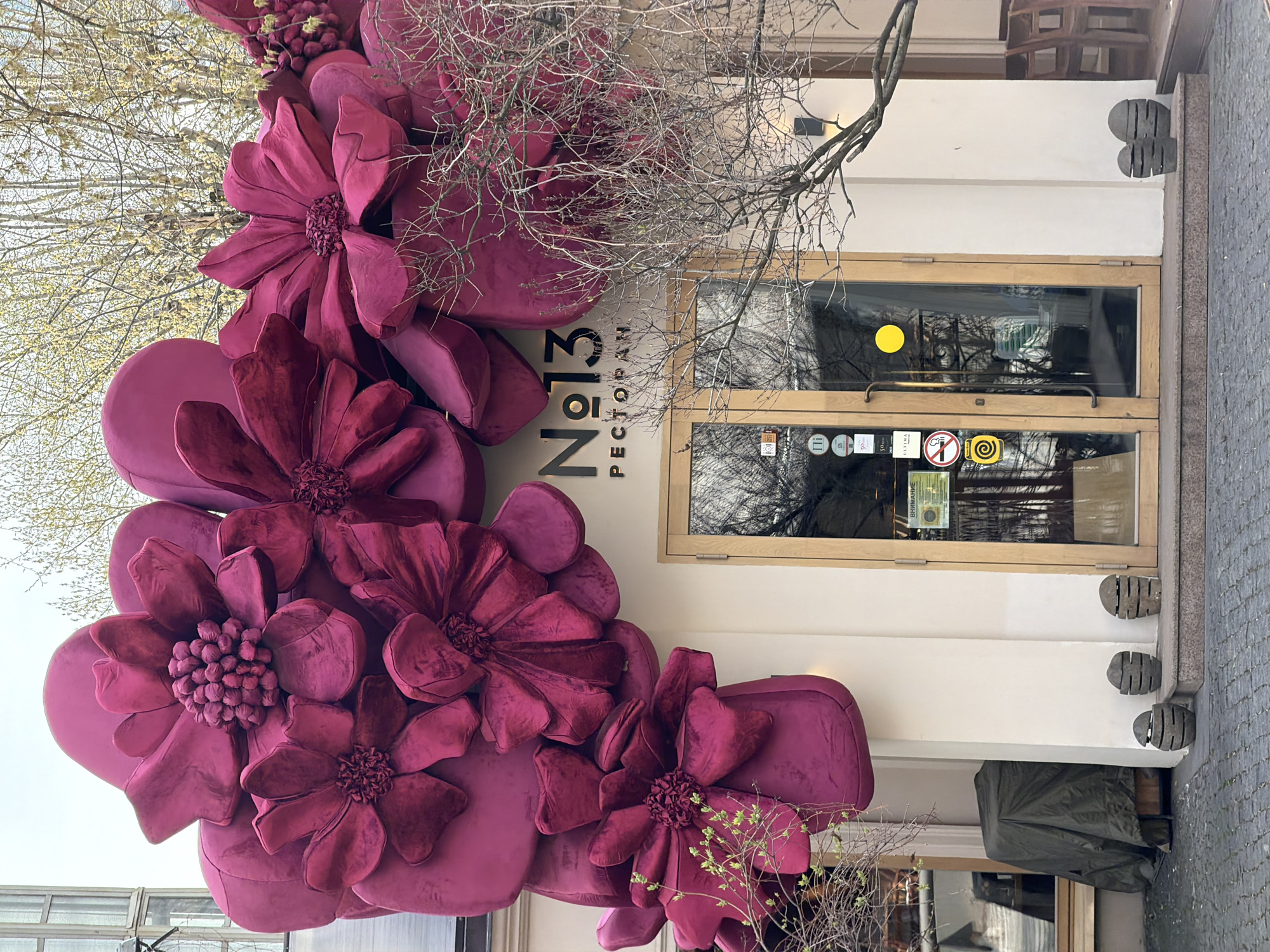 A street-level view shows the exterior of a restaurant called "No. 13 PECTOPAH" in Moscow, Russia, on a cloudy day. The facade of the restaurant is painted white with a wooden-framed glass door displaying various stickers and signage. To the left of the entrance, a large, three-dimensional floral decoration in shades of magenta adorns the wall. A leafless tree branch partially obscures the sign and decorative elements. The sidewalk and street are visible, indicating the location is an urban area.