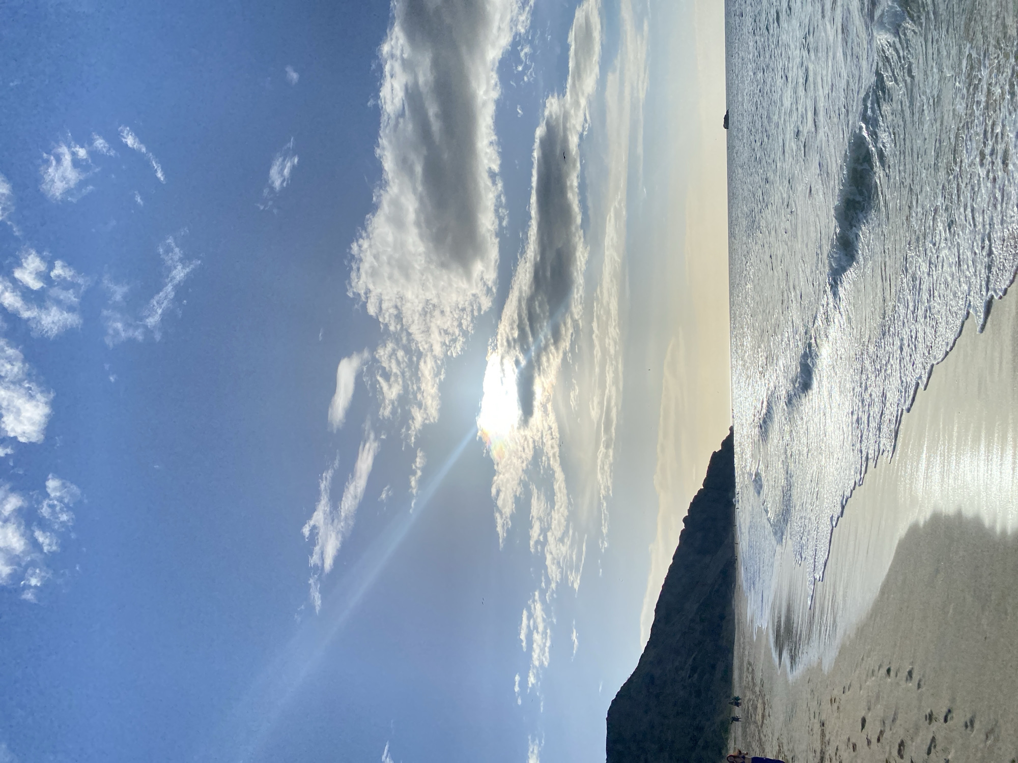 The image depicts a serene beach scene with a gently sloping sandy shore meeting the tranquil ocean. The sky is a vibrant blue, adorned with scattered white clouds, some of which are illuminated by the sun, creating lens flares and a soft glow. A distant, dark, hilly coastline forms the backdrop on the left, with a few small figures visible walking along the beach. The ocean surface is calm, with small waves lapping at the shore, reflecting the sunlight. A small, dark object, possibly a boat or a rock formation, is visible in the far distance on the water. The sand shows footprints, indicating recent human activity. The overall atmosphere is peaceful and natural. This scene does not appear to be located in Altagracia, Venezuela, as the geography does not match known locations there.