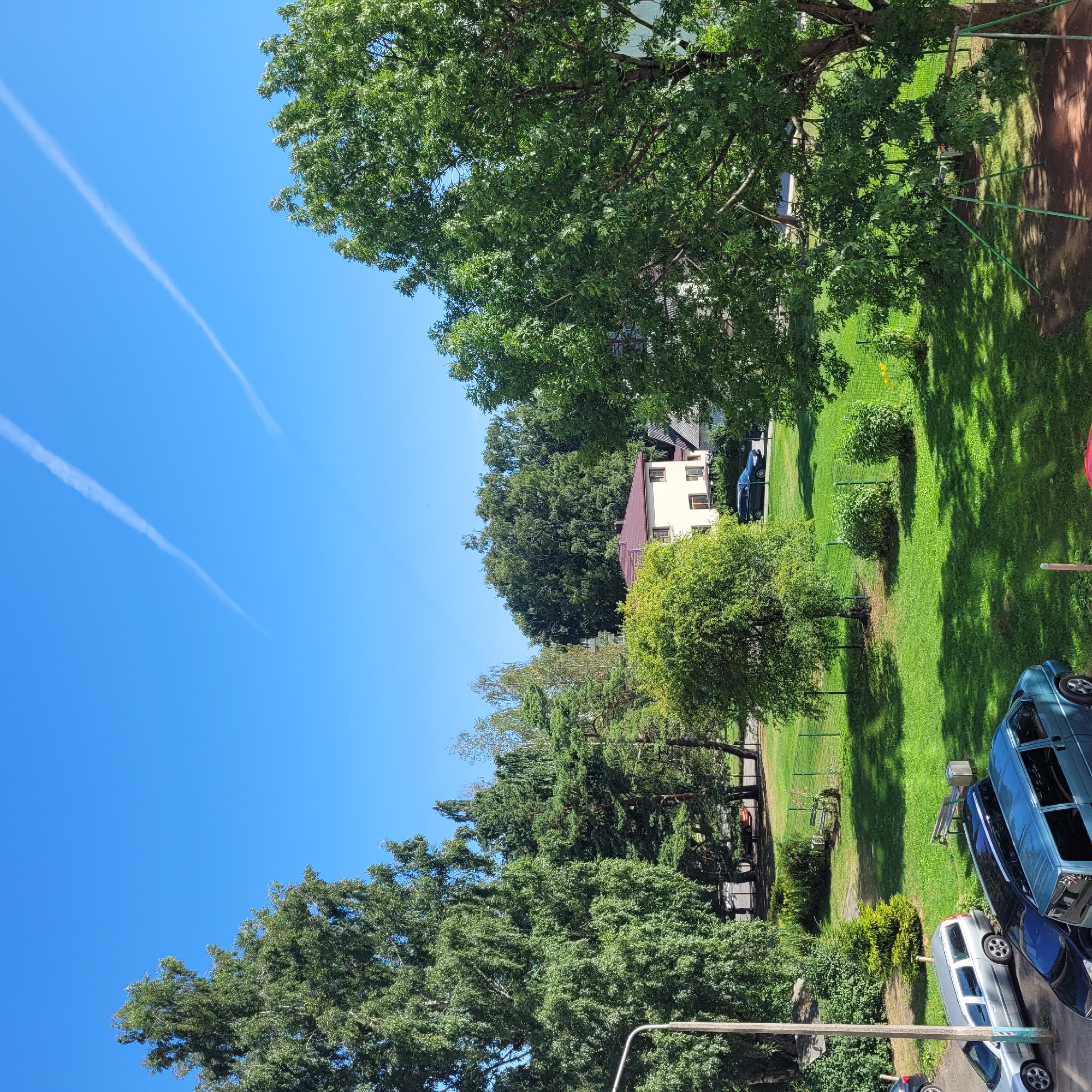The image captures a daytime scene in a residential area, likely in Jūrmala, Latvia, characterized by lush greenery and clear blue skies. A large, mature deciduous tree dominates the right side of the frame, its dense foliage casting dappled shadows across a patch of red-brown ground which appears to be part of a children's play area, complete with a green metal swing set. In the mid-ground, a neatly mown green lawn is visible, bordered by a green mesh fence. Several parked vehicles are present: a silver station wagon and a dark-colored sedan are parked on a paved area to the left, while a light blue van is parked closer to the foreground. Behind the lawn, a two-story beige house with a brown roof is situated amidst more trees. Further back, a line of diverse trees, including evergreens and deciduous varieties, forms a backdrop. The sky is bright blue with two distinct contrails arcing across it, indicating airplane activity. The overall impression is one of a peaceful, suburban environment on a sunny day.